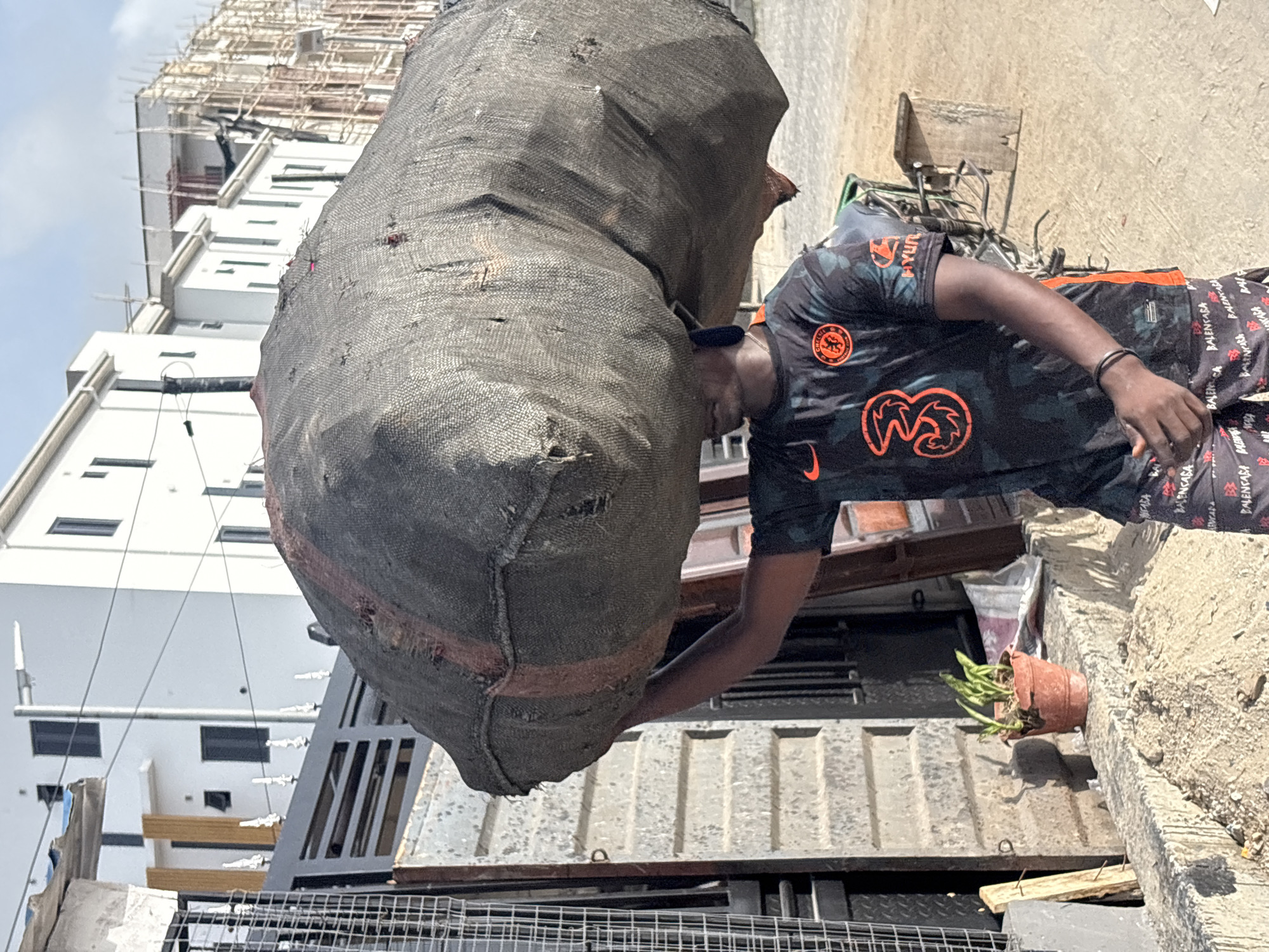 A man in a black and orange Chelsea soccer jersey and Balenciaga shorts carries a large, heavy sack over his head in Igboefon, Nigeria. He supports the sack with his left hand. Behind him are a white building, a metal gate, a rusty metal container, a plant in a terracotta pot, and a bicycle. The sky is visible in the background. A building under construction is also visible in the background.