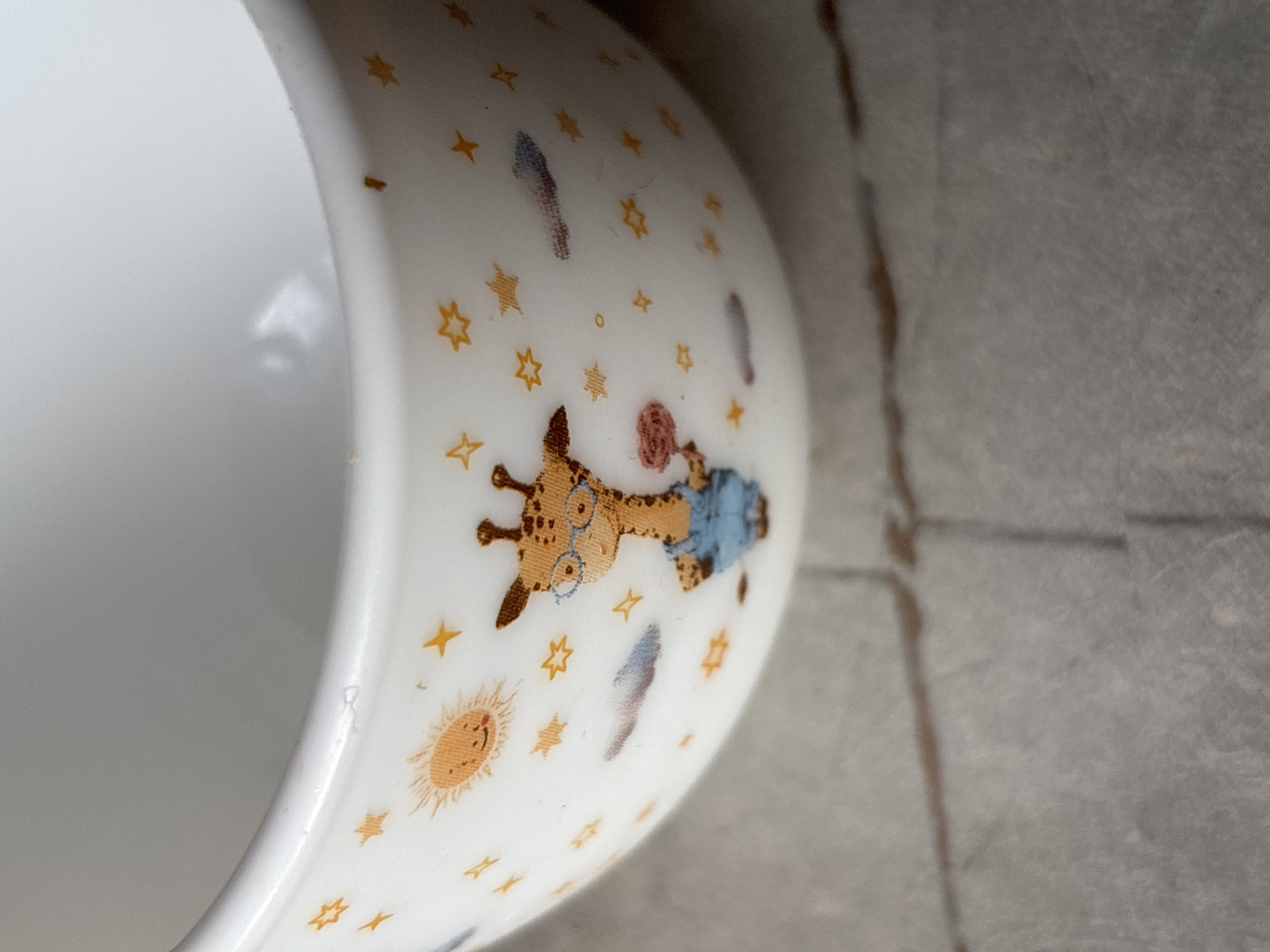 This is a close-up, eye-level shot of a white ceramic mug with a cartoon giraffe design. The giraffe is wearing glasses and blue overalls and is holding a pink object. The mug is adorned with stars and a smiling sun. The background is blurred and appears to be a gray, textured surface, possibly a tabletop or floor, with visible grout lines. The image does not contain any information about the city of Imeni Morozova, Russia.