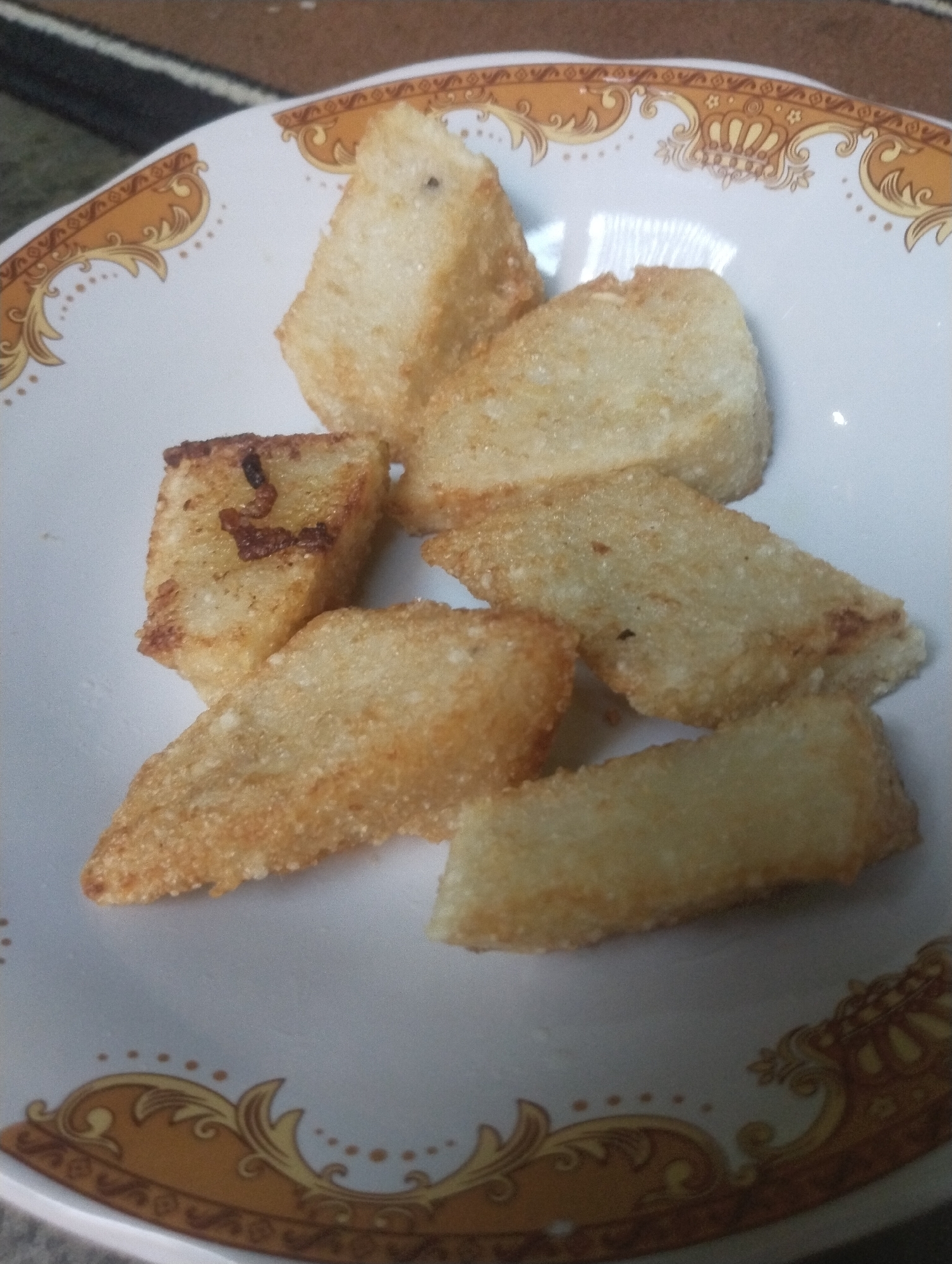 A close-up, overhead shot shows a plate of fried, rectangular pieces of food arranged on a white plate with a decorative gold and brown border. The food items appear to be a type of fried bread or cake, with a slightly golden-brown, crispy exterior and a soft interior. Some pieces are thicker than others, and the texture suggests they have been deep-fried. The plate is on a patterned surface, possibly a tablecloth or placemat, with colors of brown and gray. The image is taken in natural lighting conditions, and the focus is on the food. The location is not discernible from the image itself, and thus, the provided location of Wanasaba Daya, Indonesia is not supported by the visual evidence.