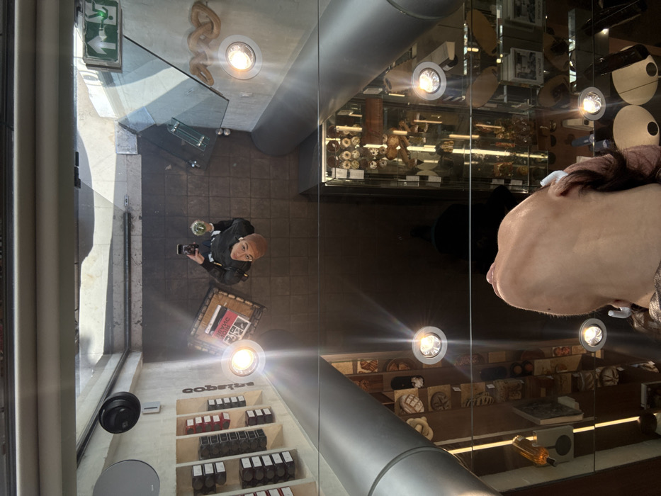 The image provides an overhead perspective through a glass floor, capturing a scene on a lower level and a reflection of the photographer above. The location is identified as the former Colette concept store, previously located at 213 Rue Saint Honoré, 75001 Paris, France.
On the lower, visible level, a light-skinned individual with very short, light-colored hair is positioned centrally, looking upwards directly at the camera. They wear a dark long-sleeved top and a light-colored undershirt or collar. An earbud is visible in their left ear. In their right hand, they hold a black smartphone, seemingly taking a photo or video. Their left hand holds a transparent cup containing a green substance and ice. The floor is composed of dark, square tiles. To the individual's left, a wooden crate labeled "POUR L'HOMAGE" (partially obscured) rests on the floor. Further left, a vertical white display shelf showcases multiple rows of boxed items, some with red labels and others with black/white labels; the word "COLETTE" is visible vertically along the shelf's edge. Above this shelf, a black dome-shaped security camera and a circular metallic vent are mounted. To the individual's right, a green emergency exit sign with a white running figure and right-pointing arrow is affixed to a grey wall. Below this sign, a coiled, serpentine-shaped decorative element is visible on the wall. Numerous bright, recessed spotlights illuminate this level, creating strong lens flares on the glass surface.
Through the glass, revealing a deeper basement or lower floor, various retail display cases filled with diverse merchandise, including small round objects and other curios, are visible. A large, grey, cylindrical pillar stands prominently on the left side of this lower level.
In the immediate foreground, a reflection of the photographer's head and shoulders is visible, looking upwards. This individual has medium-length brown hair.