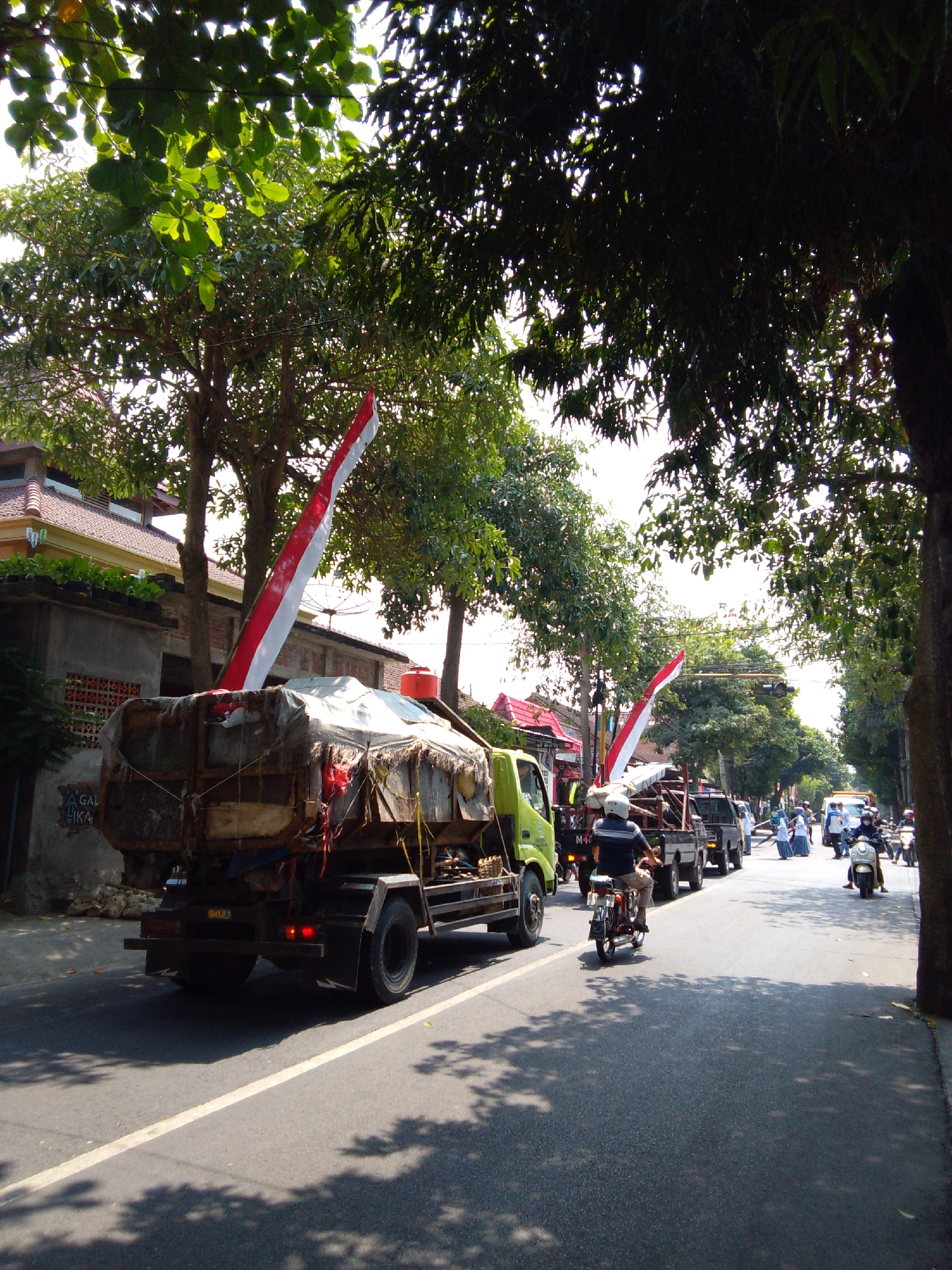 The image depicts a street scene in Trenggalek, Indonesia, on a sunny day with dappled sunlight filtering through trees. In the foreground, a large truck with a covered load is parked or moving slowly. It has a red and white Indonesian flag mounted on its side. A man in a helmet and blue shirt is riding a motorcycle behind the truck, his back to the camera. Further down the street, other vehicles, including cars and motorcycles, are visible. Several people in white clothing, possibly students or participating in an event, are standing or walking on the side of the road. The architecture visible includes buildings with red tile roofs and some with concrete facades. The overall atmosphere suggests a normal day with some organized activity or a parade element due to the flag and the gathering of people.
