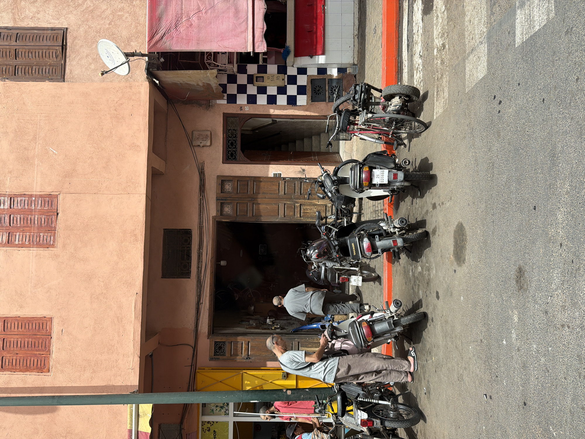 In Marrakesh, Morocco, a street scene unfolds with a row of parked motorcycles and bicycles in front of a storefront. The building's facade is painted a pale peach color, with brown wooden shutters on the upper windows. A dark green lamppost stands on the left side of the image. Below the upper windows, the building recedes, revealing an open doorway with a dark interior.
Several motorcycles are parked in a line. The closest motorcycle on the left is black with a silver trim. To its right, another motorcycle, also black, displays a license plate with the characters "26 020888". Next to it is a red and black bicycle with a cargo rack. Further down the line, more motorcycles are visible.
Two men are standing near the storefront. One man, wearing a gray t-shirt and beige pants, is leaning over a motorcycle, appearing to interact with it. Another man, dressed in a gray t-shirt and pants, is walking into the storefront. A third man, wearing a red shirt, stands further to the left near a yellow entrance.
The ground in front of the building is paved with asphalt, showing some water stains and debris. A bright orange border runs along the edge of the sidewalk, separating it from the street. The scene is illuminated by daylight, casting shadows on the ground. A satellite dish is mounted on the wall of the building on the right. The overall impression is a bustling urban environment typical of Marrakesh.