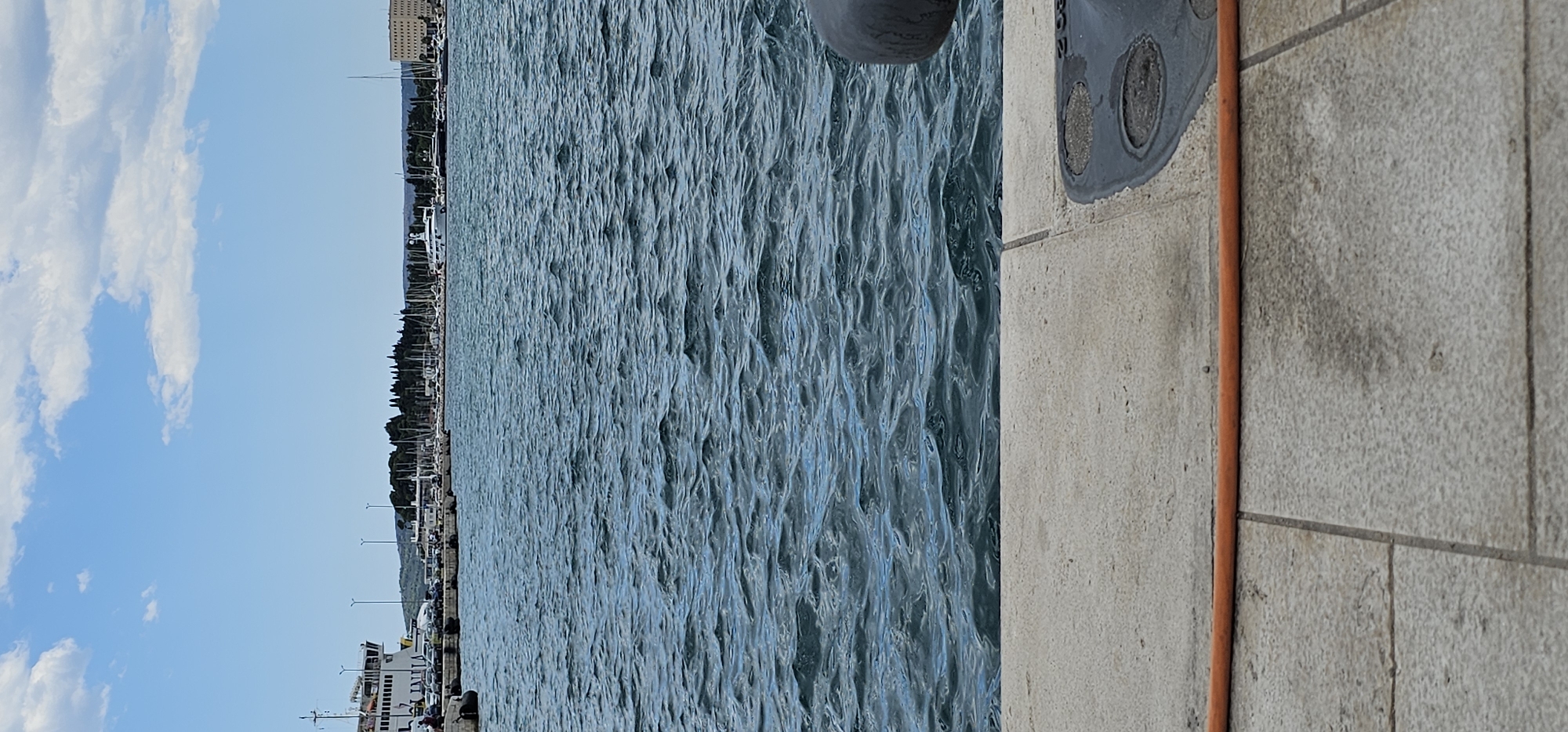 The image depicts a view of the Adriatic Sea from a harbor in Split, Croatia. In the foreground, there is a stone walkway with an orange hose lying across it. The sea is choppy with small waves. In the background, a ferryboat is docked on the left, and beyond that, a marina filled with sailboats and yachts is visible. A tree-lined embankment separates the marina from a cluster of buildings on the horizon. The sky is blue with scattered white clouds.