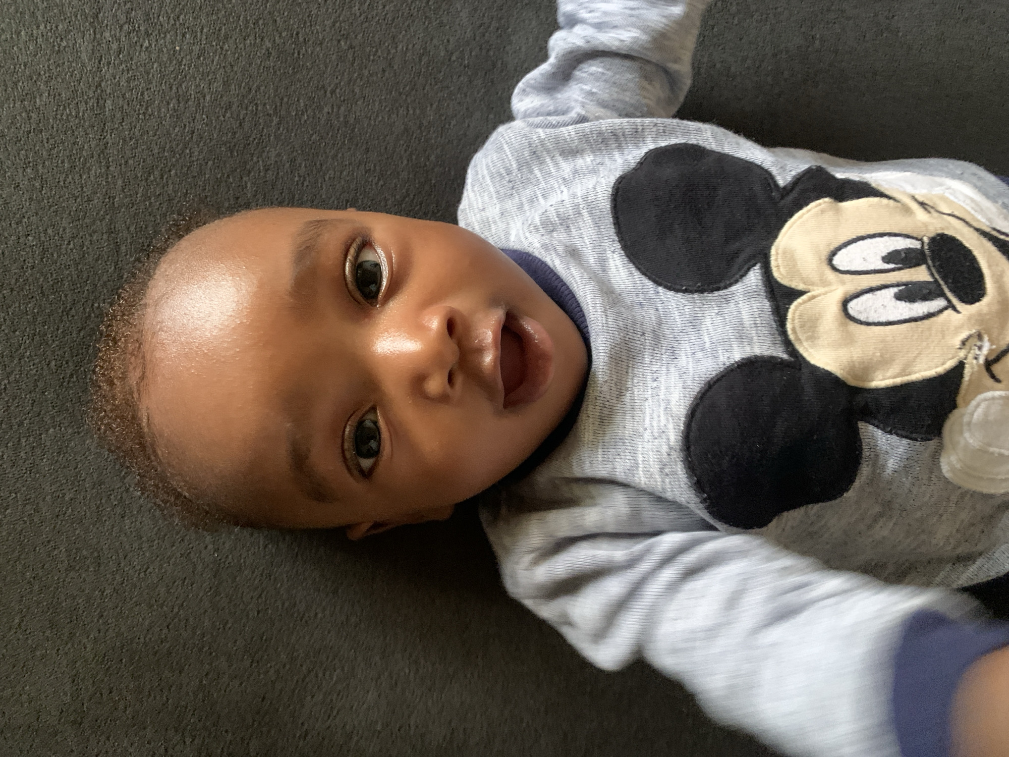 A baby with brown skin is lying on a gray surface wearing a gray long-sleeve shirt with a Mickey Mouse design. The baby is looking up and has its mouth open. The location is unknown, and there is no indication the image was taken in Barakin Akawo, Nigeria.