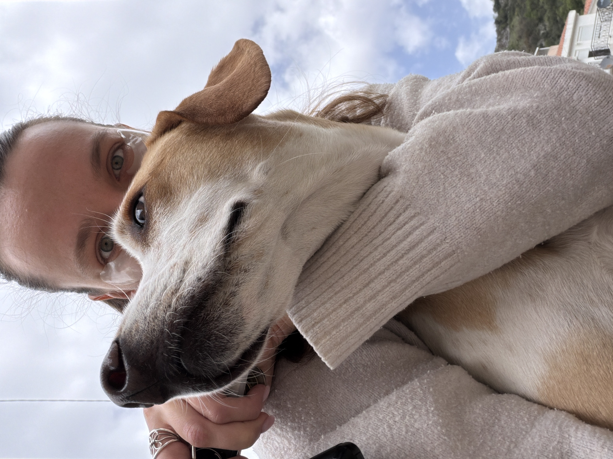 A person wearing a beige sweater is embracing a tan-and-white dog in an outdoor setting. The person has fair skin, light-colored eyes, and dark hair pulled back. The dog is in the person's arms, its head close to the camera. In the background, a cloudy sky is visible along with glimpses of buildings and a rocky hillside suggesting a coastal urban area. The exact location in Budva, Montenegro cannot be determined from this image alone.