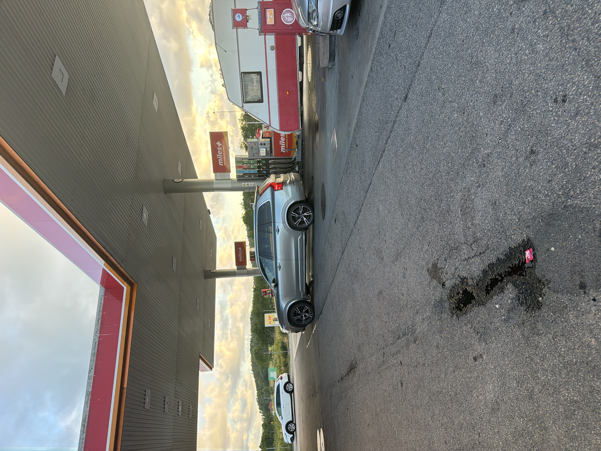 The image depicts a gas station in Hogstorp, Sweden, under a cloudy sky. The foreground is an asphalt-paved area, showing some oil stains and debris, including a red wrapper. A silver Volvo SUV is parked at a gas pump under a red and white canopy. To the right of the Volvo, a white and red caravan is parked, also near a gas pump. Further to the left, a white sedan is visible, parked at a distance. Several "Miles+" signs are present, indicating the gas station brand. In the background, there are trees and hills, suggesting a rural or suburban setting. The lighting indicates it is either early morning or late afternoon. No people are visible in the scene.
