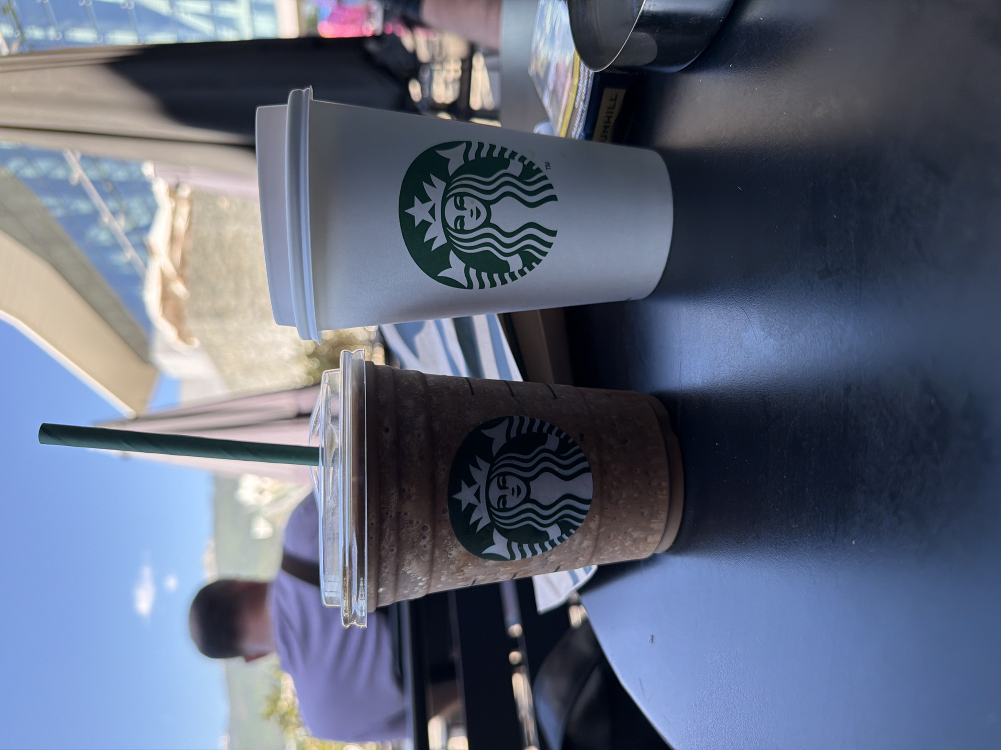 The image shows two Starbucks beverages on a dark table, likely outdoors. One beverage is a white paper cup with a green Starbucks logo, presumably hot. The other is a clear plastic cup with a green Starbucks logo, containing a blended iced drink with a green straw. A pack of Dunhill cigarettes and a metal ashtray are also on the table. Part of a person is visible in the background, wearing a white shirt. The background shows buildings and trees. Based on the architectural style visible in the background, the location is potentially in the city of Sofia, Bulgaria, near a modern building with reflective glass.
