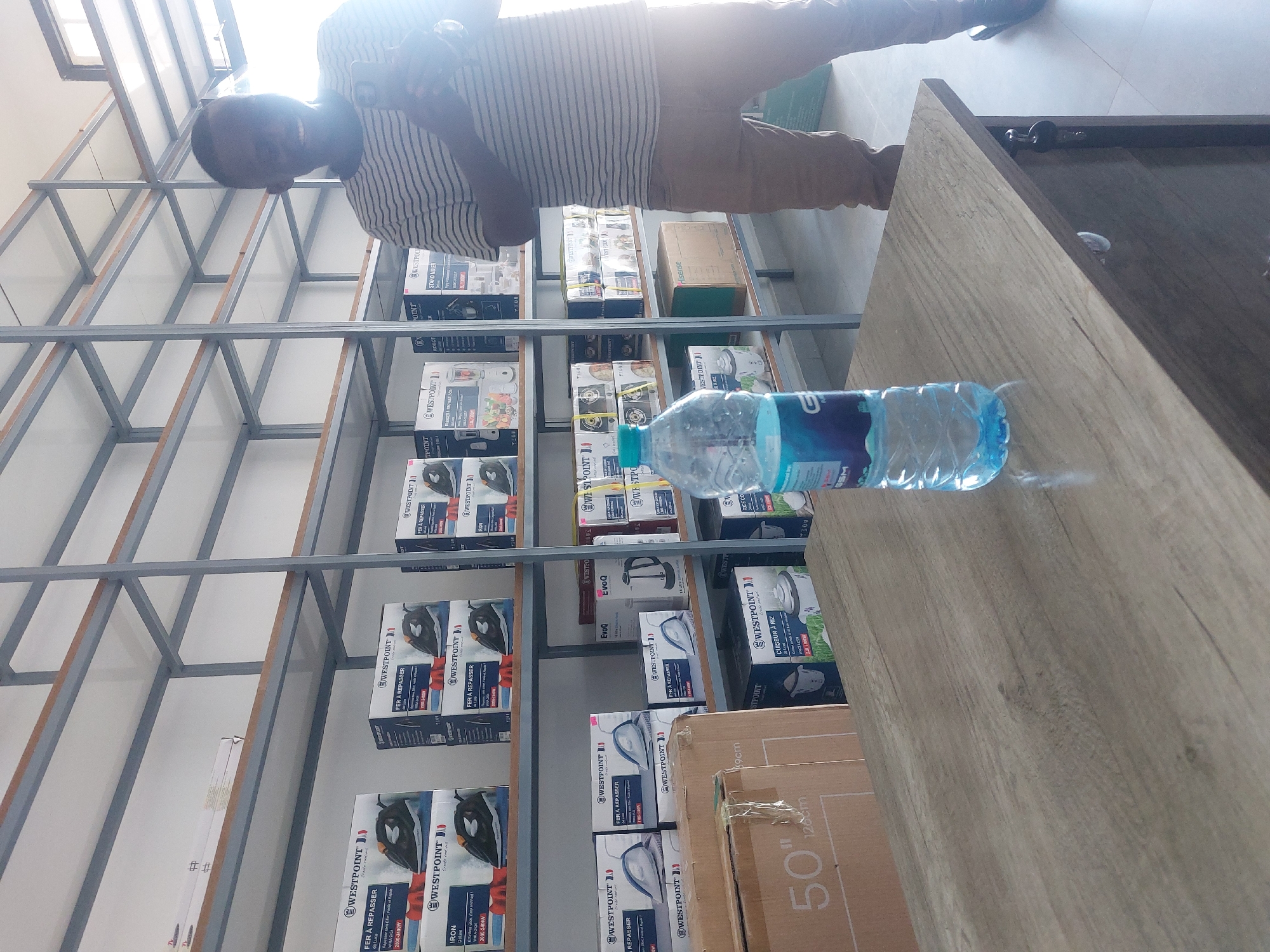 The photograph, taken indoors, presents a shelf laden with various products, including irons, blenders, and rice cookers, predominantly branded "WESTPOINT." A water bottle rests on a wooden countertop, positioned beside a cardboard box with the number "50" inscribed. The top of a man is seen holding a cellphone. The shelving unit is constructed from grey metal. Based on the visible details of "WESTPOINT" products and the general merchandise assortment, this photograph likely depicts a retail environment. The location is presumed to be Dar es-Salaam, Tanzania, but there are no corroborating details in the image to support that assertion.