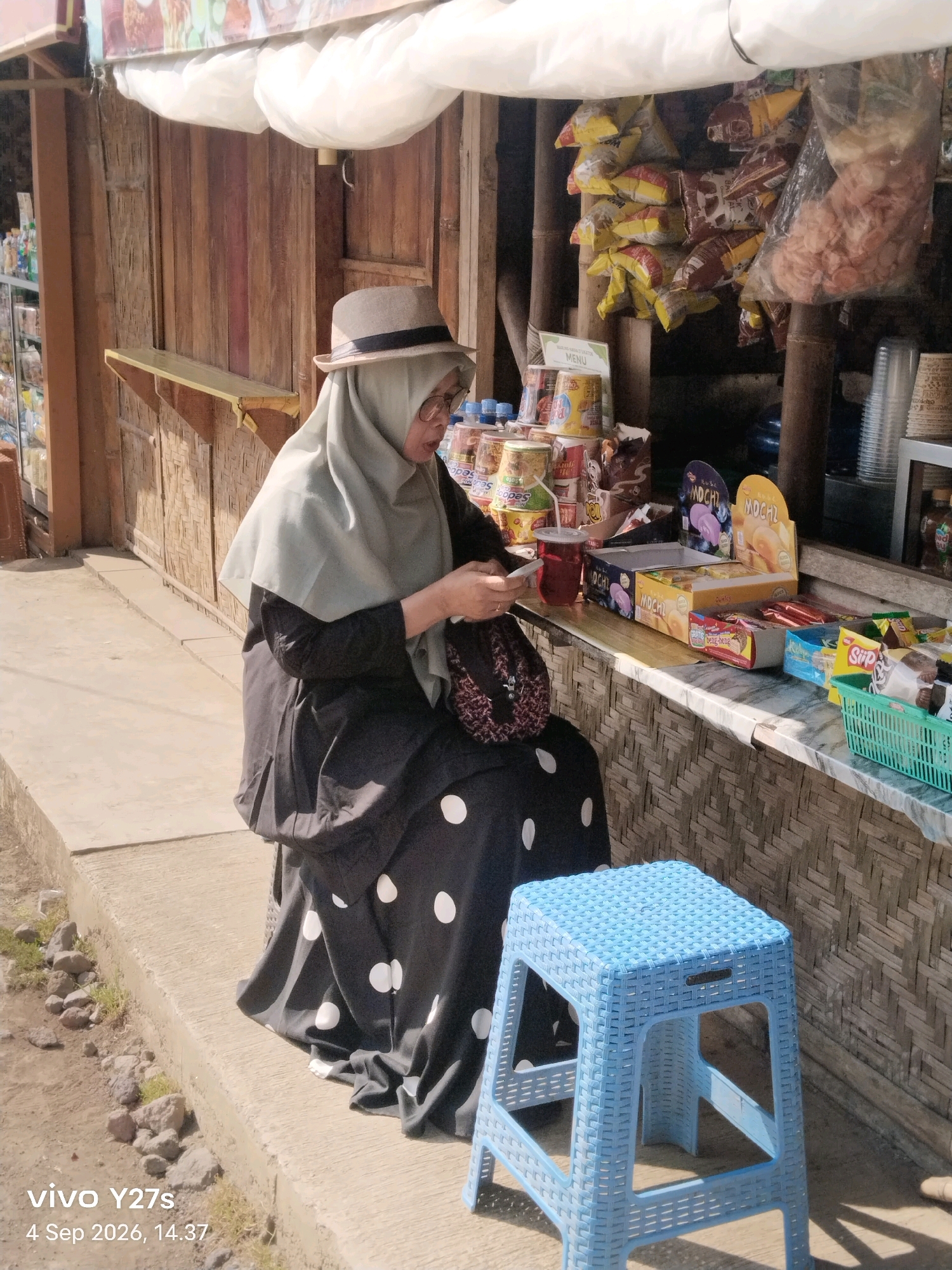 The image depicts a woman wearing a grey hijab and a black polka dot dress, sitting on a concrete ledge. She is holding a mobile phone and looking down at it. A beige hat with a dark band is perched on her head, and she is wearing glasses. In front of her, to the right, is a blue plastic stool. Behind her is a wooden stall with various products displayed, including bags of snacks and drinks. The stall has a woven bamboo backing. The ground in front of the ledge is composed of concrete and small stones. The lighting suggests it is daytime, with strong shadows cast by the stool. A watermark in the lower left corner indicates the image was taken with a vivo Y27s phone on September 4, 2026, at 14:37. The scene appears to be an outdoor vendor stall in Sigedang, Indonesia.