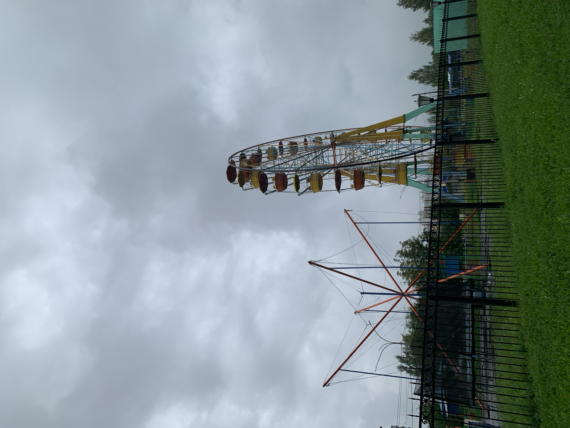The image depicts a cloudy day at an amusement park in Prokopyevsk, Russia. In the foreground, a well-maintained green lawn is bordered by a black ornamental metal fence. Behind the fence, a trampoline with elastic cords is visible, as is a section of a large Ferris wheel with colorful cabins. Trees and buildings are seen in the background, along with lampposts. The overall impression is of a quiet, overcast day in an outdoor recreational area.