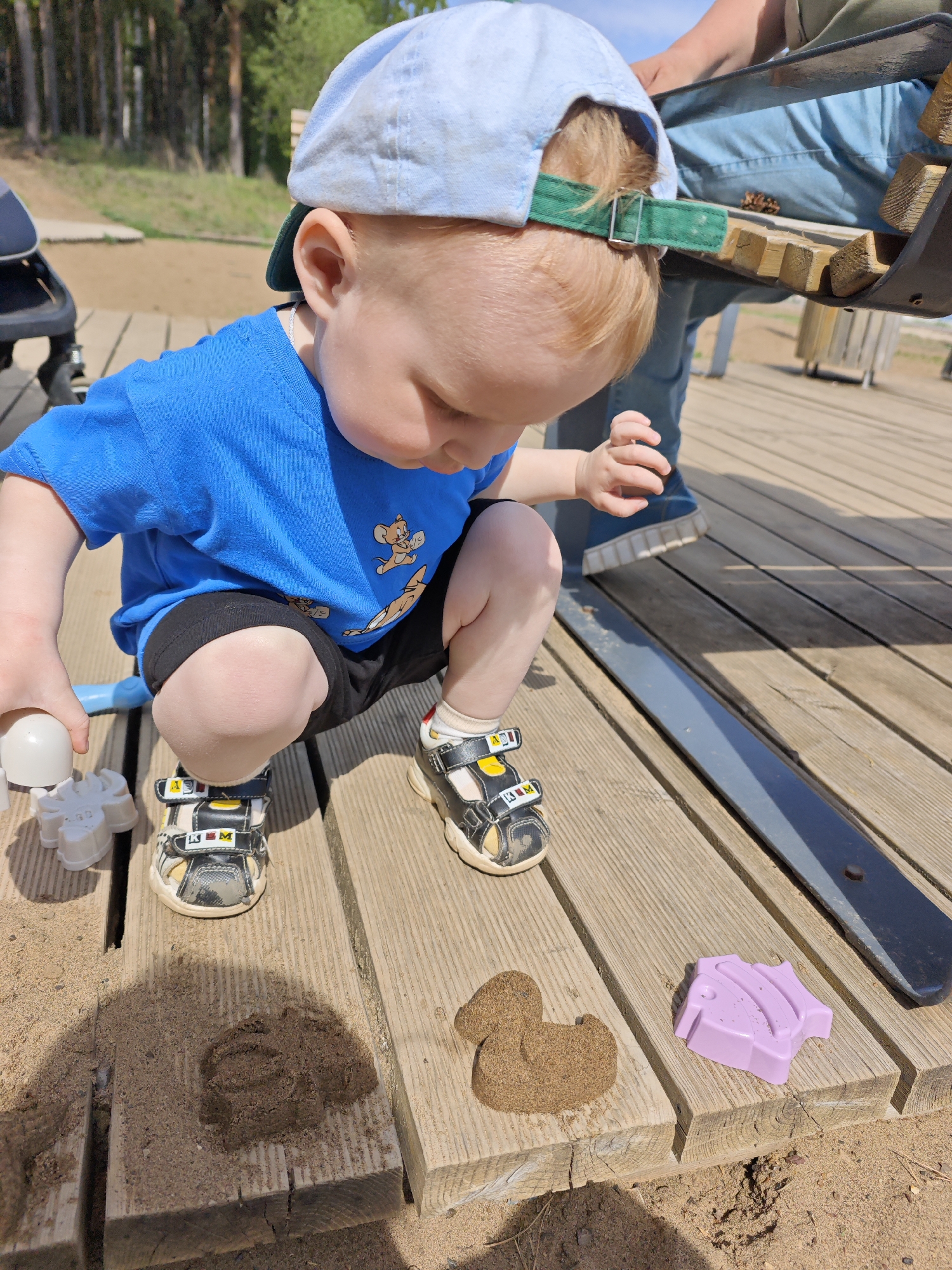 A young child is squatting on a wooden boardwalk at a playground, manipulating sand with toys in the city of Chaikovsky, Russia. The child, wearing a blue t-shirt with cartoon characters, black shorts, and sandals, is also wearing a light blue baseball cap with a green brim. The child holds a white toy in one hand and is interacting with sand formations on the wooden planks. A brown, sand-formed duck shape and a lavender, plastic toy are visible on the boardwalk near the child. A portion of a wooden bench is visible behind the child, along with an adult's legs and shoes. The background includes trees, suggesting a park or outdoor setting. Sunlight indicates it's daytime.