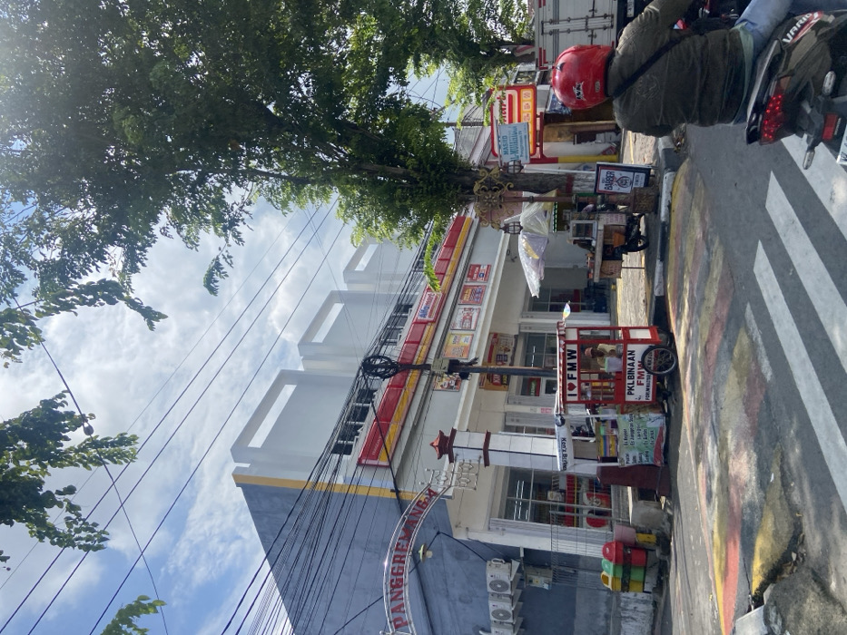 The image shows a street scene in Mojokerto, Indonesia, at the coordinates -7.478 latitude and 112.44 longitude. A large, multi-story building with a red and yellow awning is visible on the left side of the frame. The building is adorned with various signs and banners, including the word "PANGGREMANA" written in large letters.
The street is paved with asphalt and has a crosswalk. A red and white street lamp is positioned at the edge of the sidewalk.
On the right side of the frame, a person wearing a red helmet is riding a motorcycle. The motorcycle is parked in front of a small building with a red sign.
In the foreground, there are several street vendors selling their wares, including a fruit vendor and a food vendor.
The sky is partly cloudy, and there are power lines running overhead. A large tree with green leaves is on the right side of the frame.