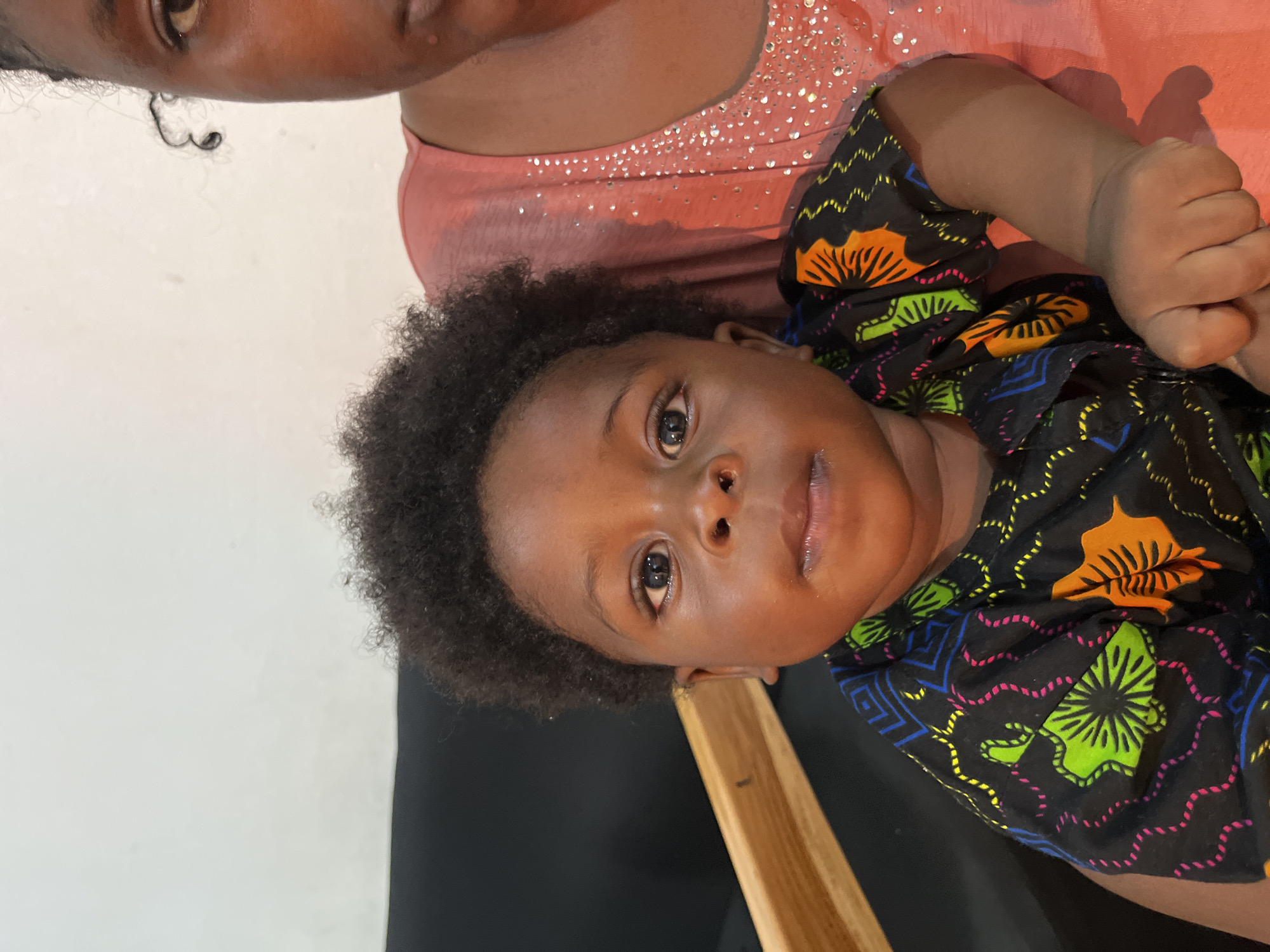 A close-up, eye-level, outdoor portrait shows a dark-skinned baby being held by an adult. The baby, facing forward and looking directly at the camera, has dark curly hair and is wearing a black shirt with a colorful African print. The adult, whose face is partially visible on the right side of the frame, is wearing a coral-colored shirt adorned with sequins. The background consists of a white wall and a dark surface, possibly a couch, with a wooden beam visible behind the baby. The scene is likely captured in Gura Top, Nigeria, given the context.