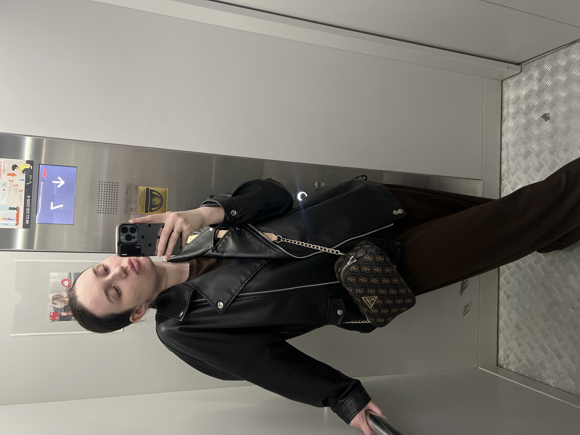 A young woman is taking a selfie in an elevator. She is wearing a black leather jacket, brown clothing, and a small patterned purse with a gold chain. She is holding a phone in her right hand. The elevator has a metal control panel with buttons and signage in Russian. The interior walls are white and the floor is metallic. The picture suggests that this image was taken in Tyumen, Russia, as the signage is in Russian.