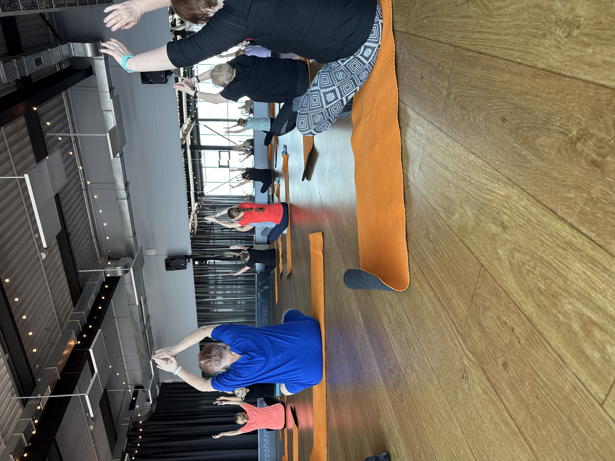 A group of people are engaged in a yoga class inside a spacious room with a high ceiling. The room appears to be located in Votkinsk, Russia, based on the partially visible word "ПОБЕД" on a mat, which likely indicates the street name "ул. Победы" (Victory street) that is located in Votkinsk. Participants are positioned on orange yoga mats, performing an arm-stretching exercise. Most participants are older adults. The room is well-lit with both natural light from the windows and artificial light from overhead fixtures. A black curtain is partially visible at the far end of the room.