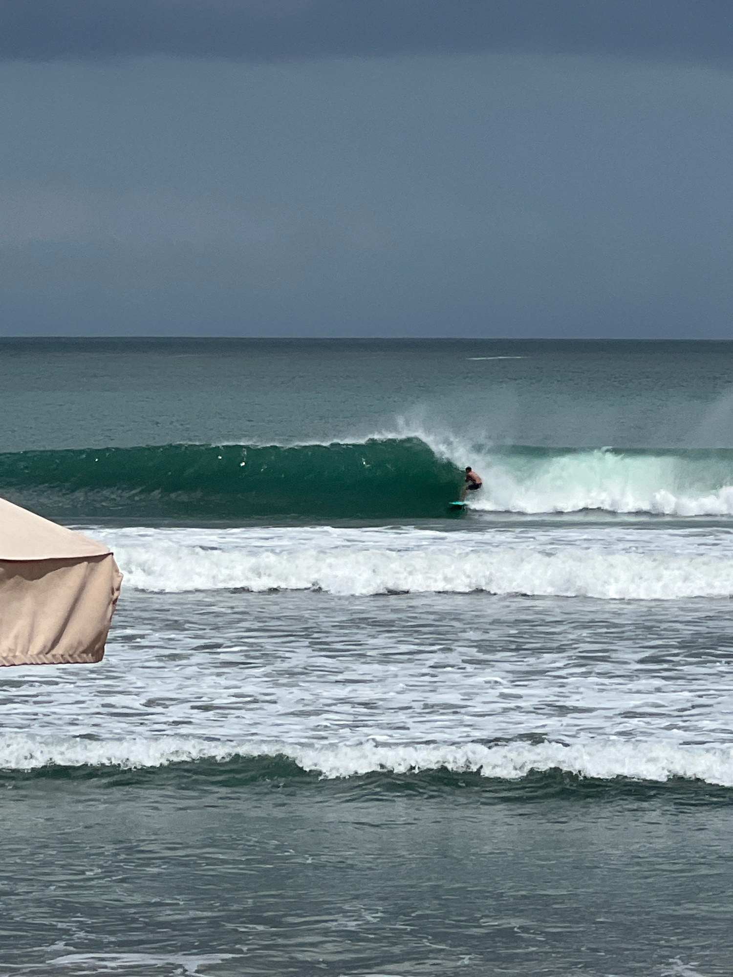 A surfer rides a green wave in Kuta, Indonesia. The surfer, seen from the side and from the chest up, is positioned in the center of the frame on a turquoise surfboard. The wave is breaking in the mid-ground, with white foam and spray erupting around it. The ocean in the foreground is choppy, with several smaller waves breaking. A section of a large, beige umbrella is visible in the lower left corner of the frame. The sky above is dark and overcast, with a slight gradient from dark gray at the top to a lighter gray near the horizon. The horizon line is visible between the sky and the sea.