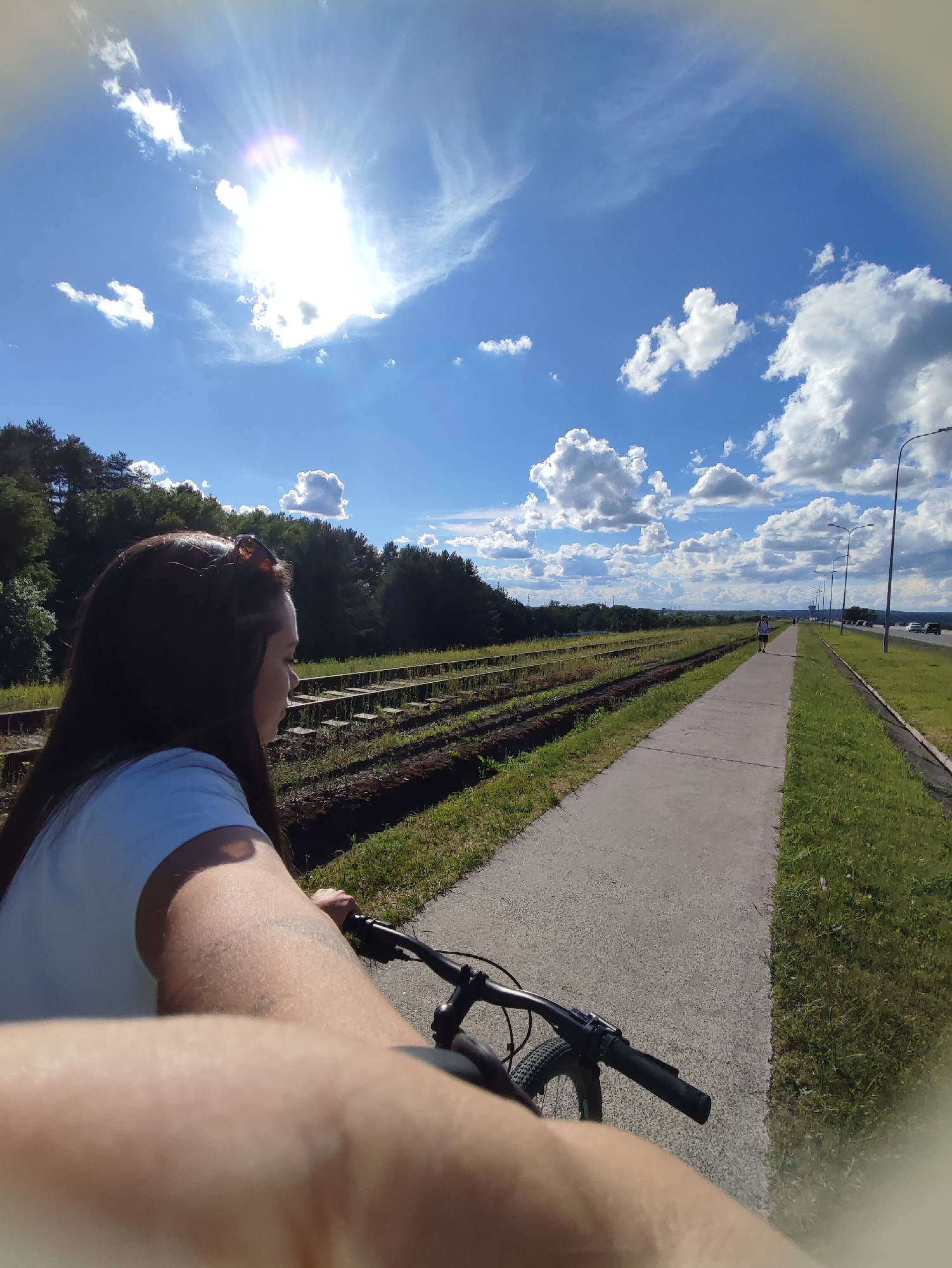 The image depicts a first-person perspective of a person riding a bicycle along a paved pathway adjacent to a railway track and a road. The person is partially visible, showing a left arm, handlebar of the bicycle, and the back of the head. The sky is predominantly blue with scattered clouds and a bright sun. Trees are visible along the railway track. The scene appears to be outdoors during a sunny day. It shows the railway track and the bike path along Sovetskaya Street in the city of Chaikovsky, Russia. The road to the right has multiple lampposts.
