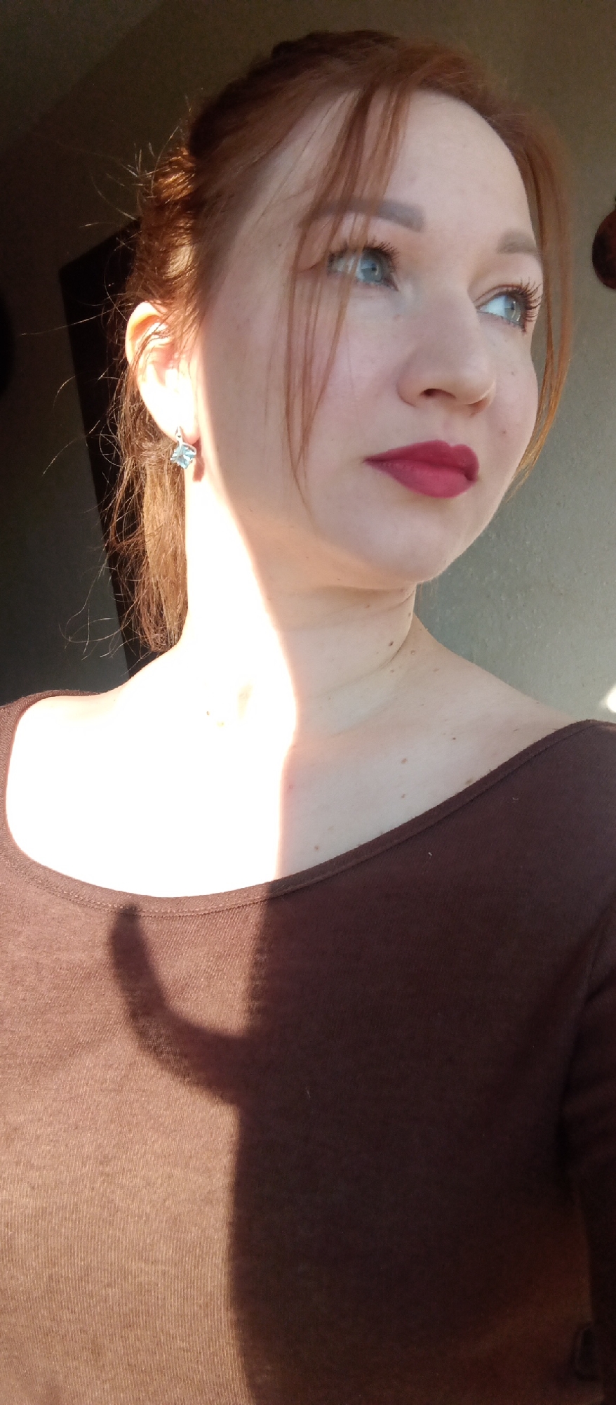 A close-up, half-body shot captures a woman in profile, looking towards the right side of the frame. Her head is tilted slightly upwards, and her gaze is directed away from the viewer. She has reddish-brown hair, styled back in what appears to be a loose bun or ponytail, with some strands falling forward around her face. Her light blue eyes are visible, along with defined eyebrows and dark mascara. She wears vibrant red lipstick.
A strong, warm light source, likely sunlight from a window, illuminates the left side of her face, her ear, and her neck, casting bright highlights on her skin. She is wearing a dark brown, long-sleeved top with a wide, boat-neck style neckline. A distinct, dark shadow is cast across her chest and shoulder, contrasting sharply with the brightly lit areas. An earring with a light-colored, possibly blue or clear, gemstone is visible on her left ear.
The background is softly blurred and appears to be an indoor setting with a light-colored wall on the right and a darker area on the left. The lighting suggests either early morning or late afternoon. There is no visible text in the image.