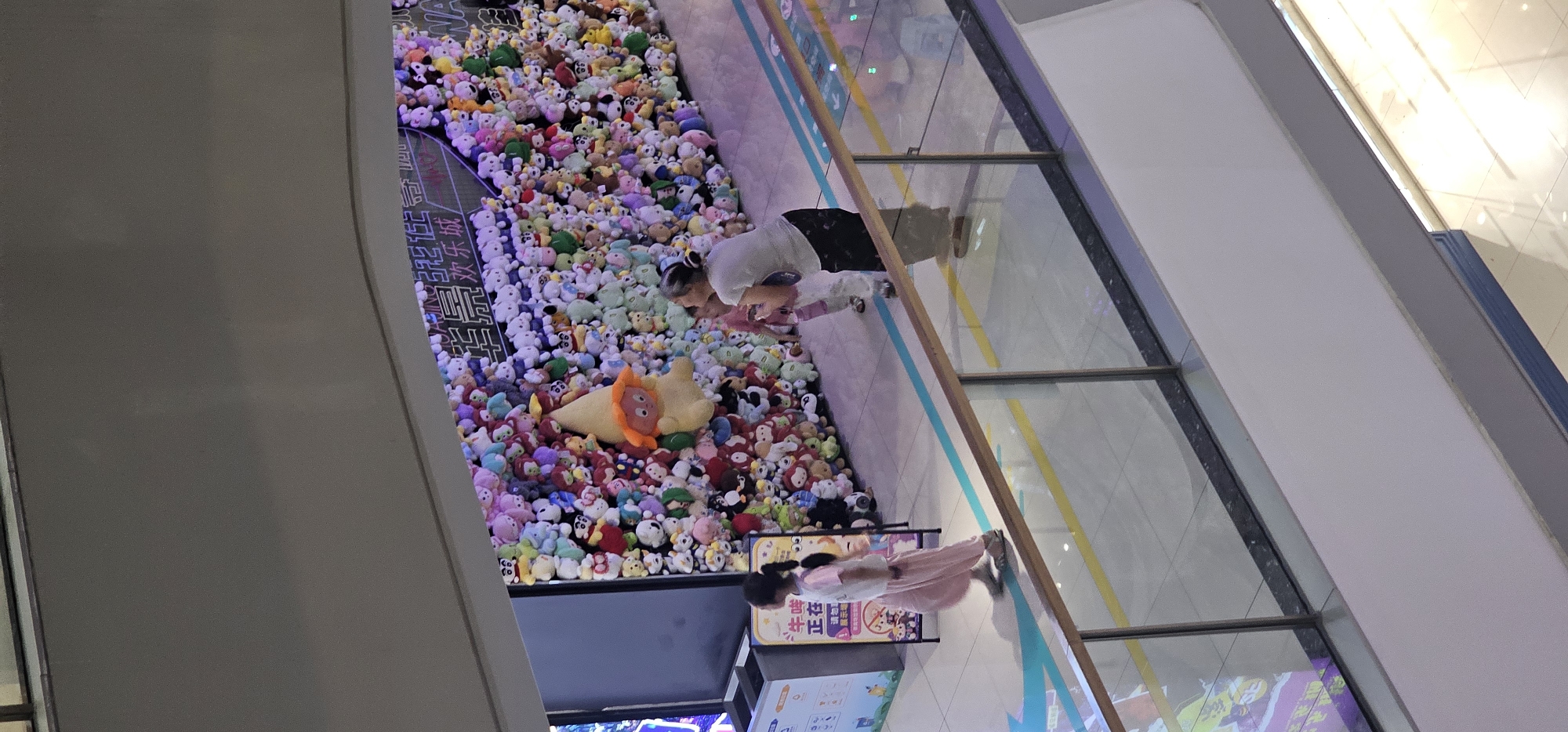This is an eye-level, medium shot photograph taken in the Shapingba District, China, showing an indoor shopping mall. The main subject is a wall covered with a multitude of plush toys, creating a vibrant and colorful display. In the foreground, a woman with her hair in a bun, wearing a white t-shirt and black patterned pants, is looking down and to the right. She is interacting with a young child standing in front of her, also looking down. To the left of the woman and child, a young girl with pigtails, wearing a pink t-shirt and loose pink pants, walks away from the camera and towards the left. She is wearing sandals. Behind the people and the plush toy wall, there appears to be a display screen or advertisement. A glass railing with a wooden top separates the viewer's perspective from the scene below. The floor is tiled, and there are colored lines on the floor, possibly indicating pathways. The lighting suggests an indoor environment, possibly with artificial lights illuminating the scene.