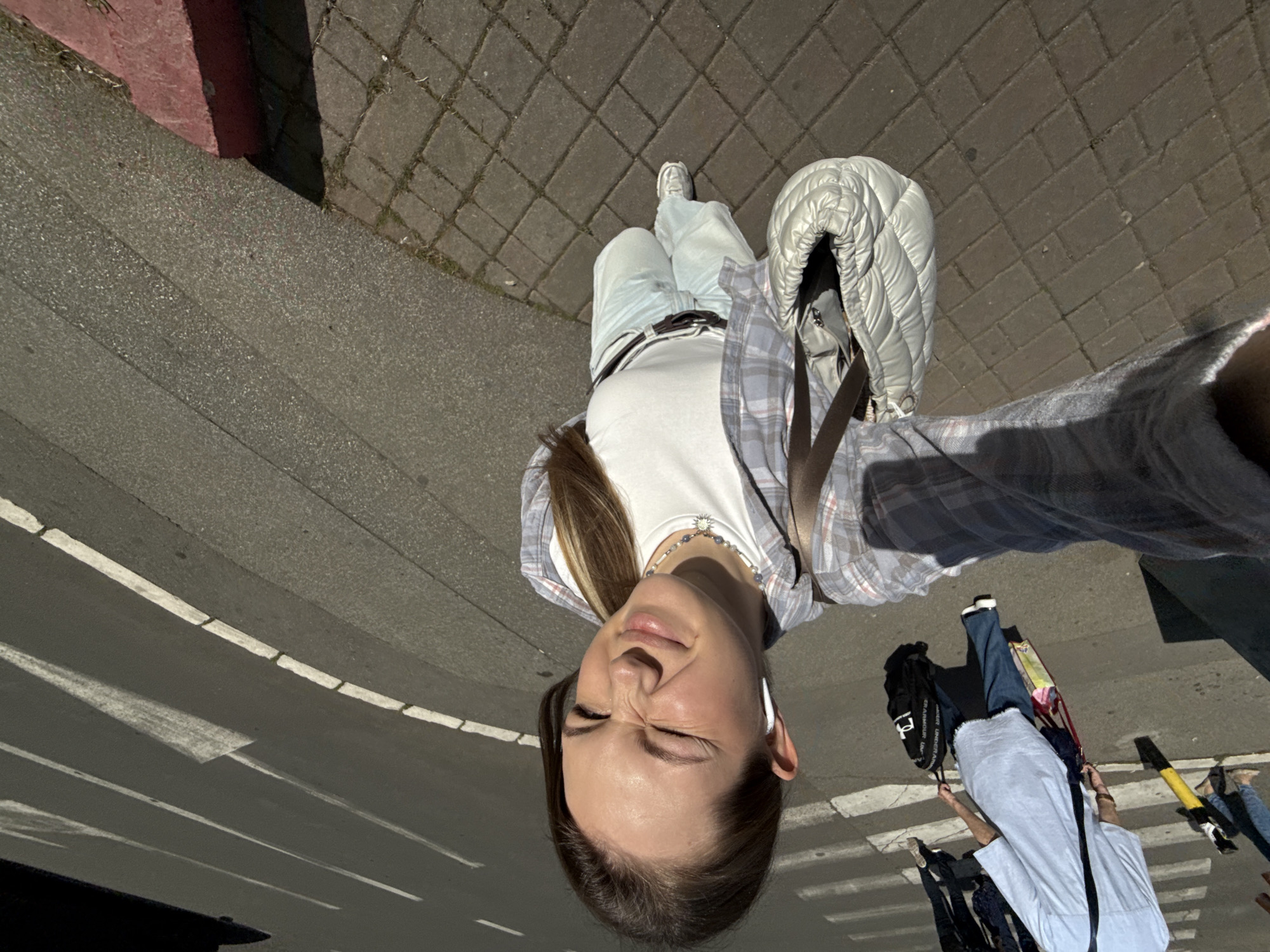 The image shows a woman taking a selfie in an urban environment. She is standing on a sidewalk adjacent to a road with white painted crosswalk markings. The sidewalk transitions into a paved area with brick-like patterns. The woman is wearing light-colored jeans, a white t-shirt, and a plaid shirt. She has a white, quilted jacket hanging from her shoulder. Other pedestrians with bags, luggage, and other belongings can be seen crossing the crosswalk. It's difficult to specifically pinpoint the exact location in Vracar Urban Municipality, Serbia from only this image. Further context is required for a forensic identification.