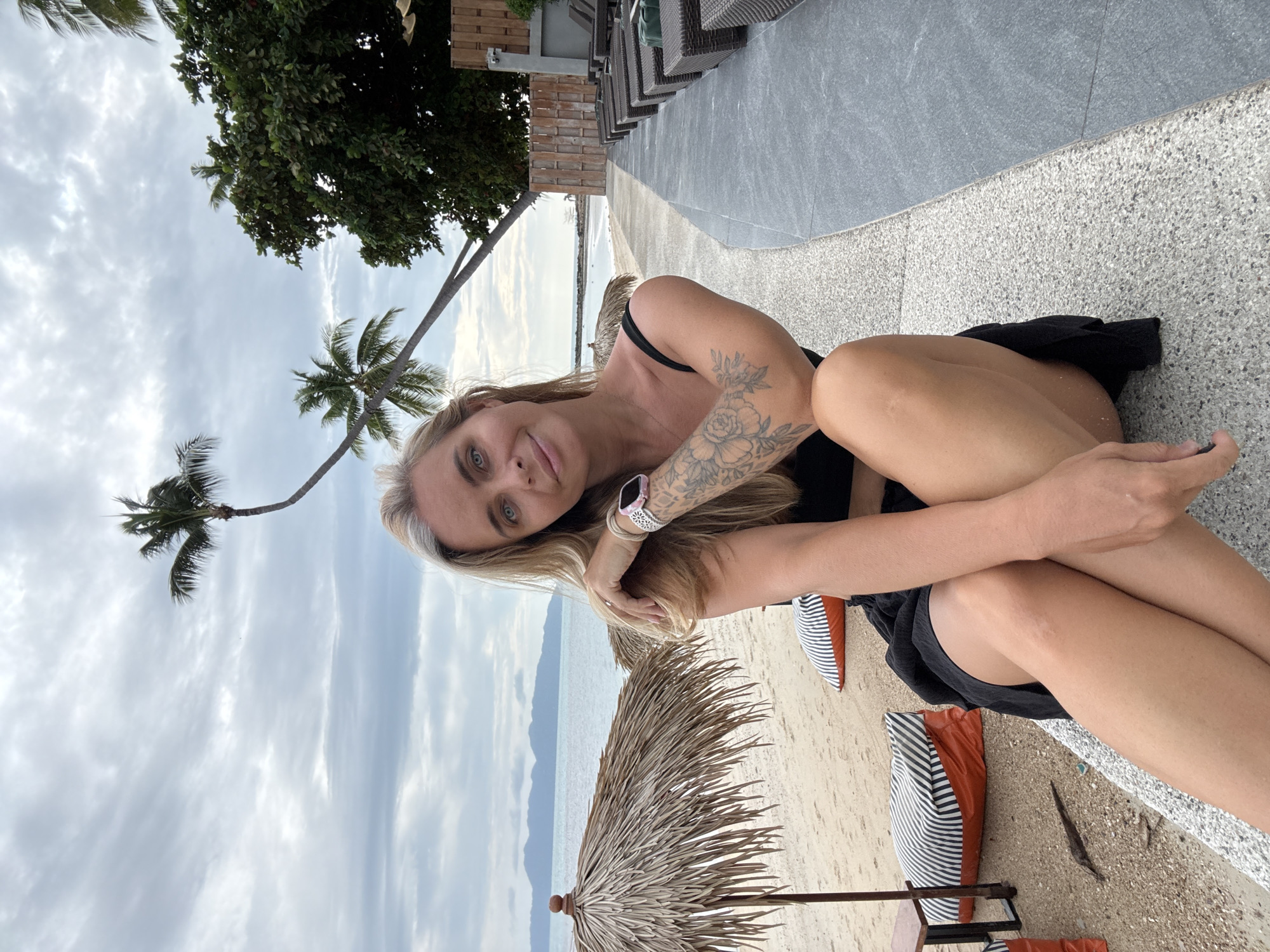 A woman with blonde hair, blue eyes, and tattoos on her left arm is sitting on a concrete ledge on a beach in the Ko Samui District of Thailand. She is wearing a black bikini top and a black skirt. The beach has white sand, palm trees, and umbrellas. In the background, there is the ocean and mountains. To the right of the woman, there are lounge chairs. The sky is cloudy.