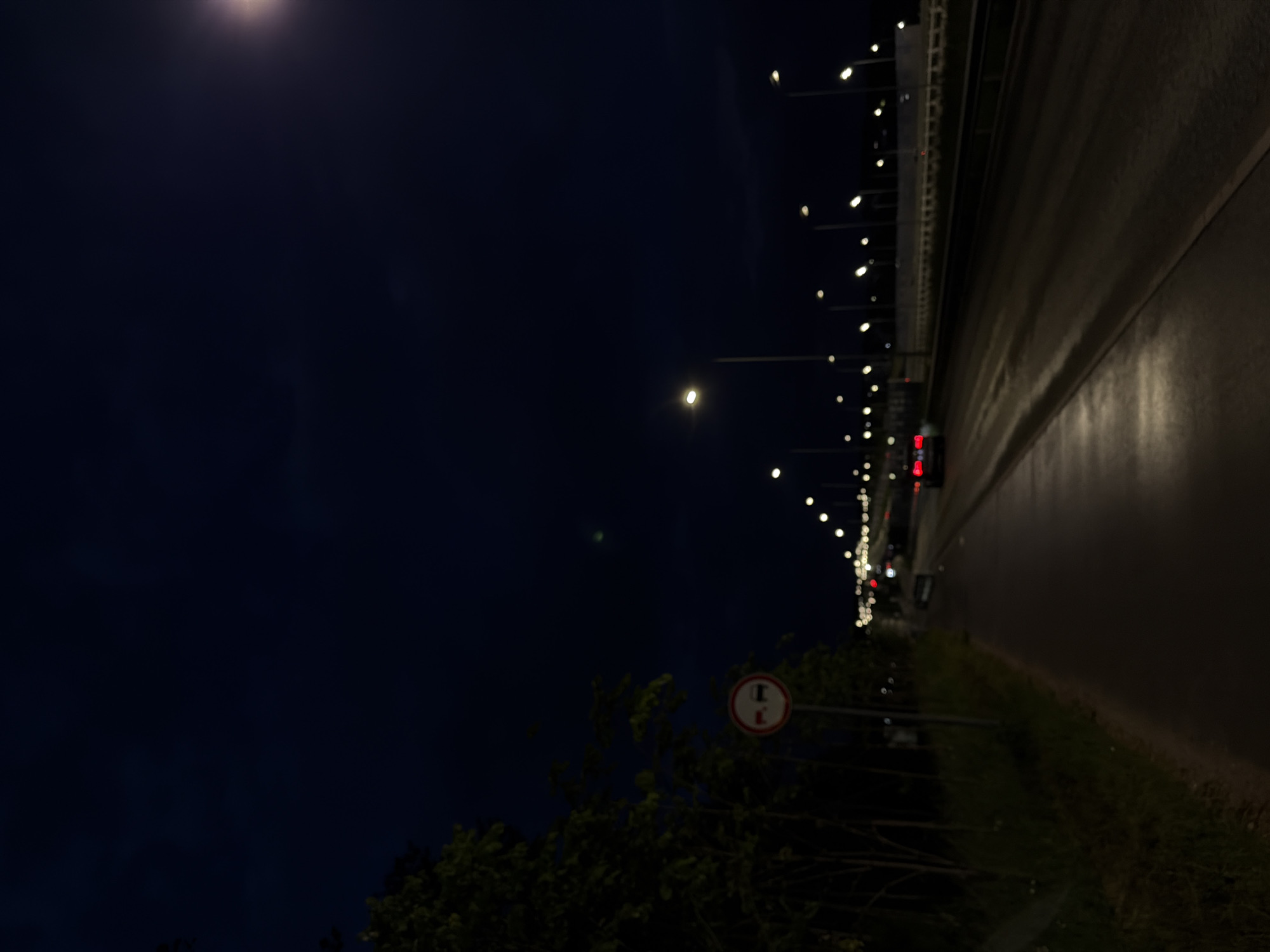 An outdoor, eye-level, medium shot shows a road at night in an unspecified city. A bright streetlight illuminates the right side of the paved road. The dark sky above has a single, blurry light source on the upper right side. To the left, a sidewalk or bike path runs alongside a line of dark trees and grass. A circular traffic sign with a red border and white background, depicting two red cars with a diagonal red bar across them, is visible on a pole on the left side of the path. A car with illuminated red taillights drives away from the viewer on the road. The road continues into the distance, lined with more streetlights and a concrete barrier.