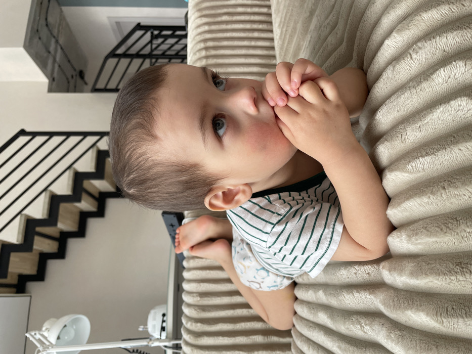 A young child is depicted lying prone on a ribbed, light-beige sofa. The child has light brown hair and blue eyes, with flushed cheeks. They wear a white short-sleeved t-shirt with thin green horizontal stripes and a light-patterned diaper or pull-up. Their bare feet are crossed behind them. Both hands are clasped near their mouth, and their gaze is directed upwards and to the right. In the background, a modern interior is visible, featuring a white desk lamp, a small white electronic device, and a dark remote control on a surface. A contemporary staircase with dark railings and light-colored treads ascends upwards against a light wall. Exposed concrete ceiling with visible conduits is also present. The specific location within Izhevsk, Russia, cannot be determined from the provided image.