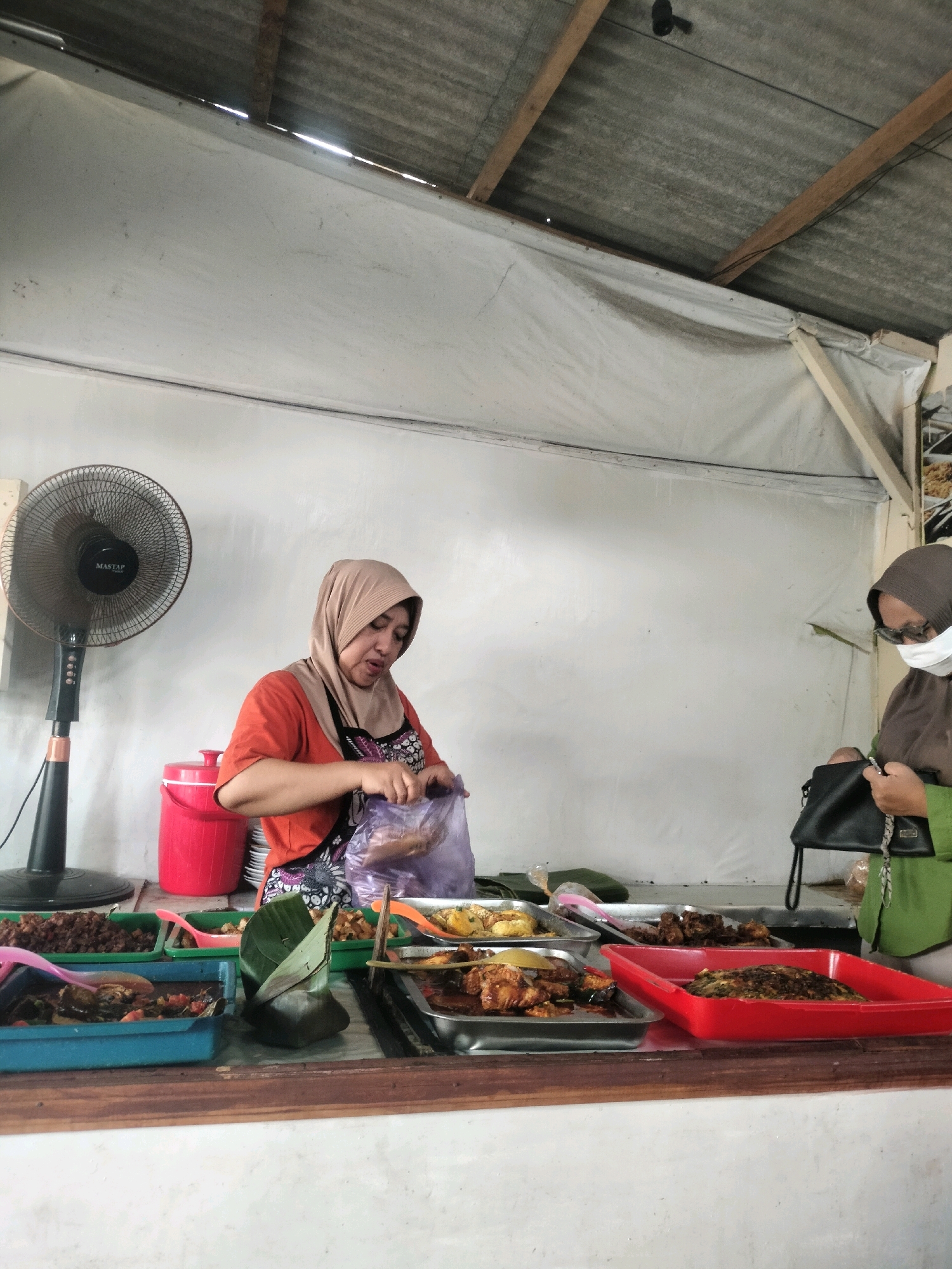 The image depicts an outdoor food stall in Tersono, Indonesia. A woman wearing a beige hijab and an orange shirt is standing behind a counter, preparing food in a plastic bag. She is wearing an apron. In front of her are various trays of food, including what appears to be fried chicken, vegetables, and rice dishes. To her left is a black oscillating fan. To her right, a woman wearing a brown hijab and a green shirt, with a white face mask, is standing with a black handbag. The stall is covered by a corrugated metal roof supported by wooden beams and white tarpaulin walls. The background is a white wall.