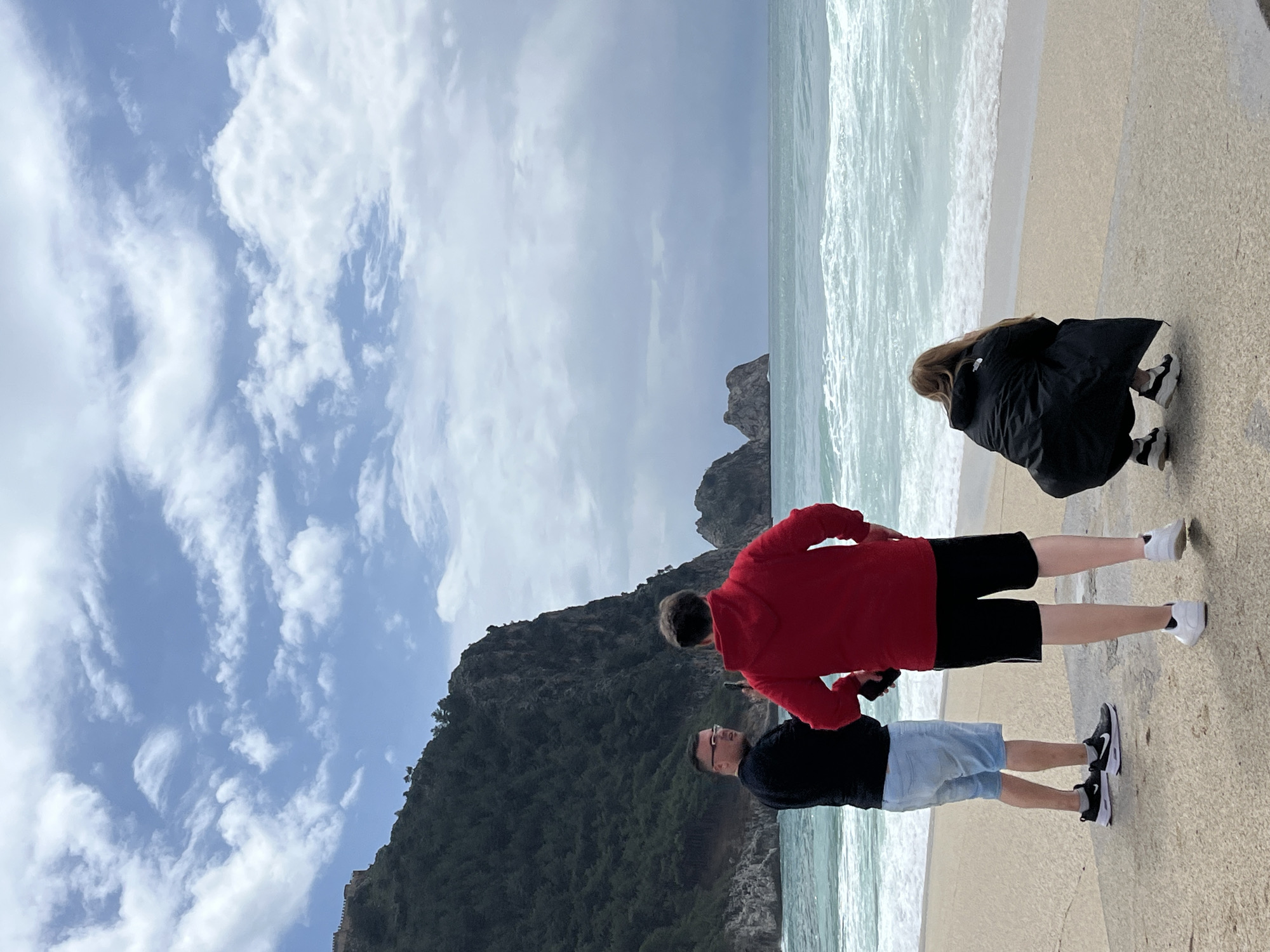On a beach in Alanya, Turkey, three people are positioned. The foreground features a man in a black shirt and light blue shorts, standing next to a man in a red sweatshirt and black shorts. In the background, a person in a black coat is crouched down. The beach leads to the ocean, where waves break near the shore. A large, rocky mountain rises in the distance. The sky is filled with clouds.