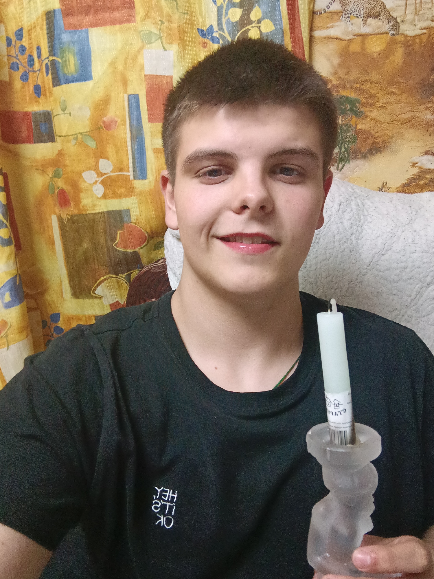 A person is holding a candle in a candle holder in an indoor environment. The person is fair-skinned, with short brown hair. They are wearing a black T-shirt with white text on it. The candle is white and has a label on it. The candle holder appears to be made of glass or crystal and is shaped like a person. The background consists of a yellow patterned curtain and a white pillow. There is no visual information in the image to suggest it is located in the city of Красный Маяк, Russia.