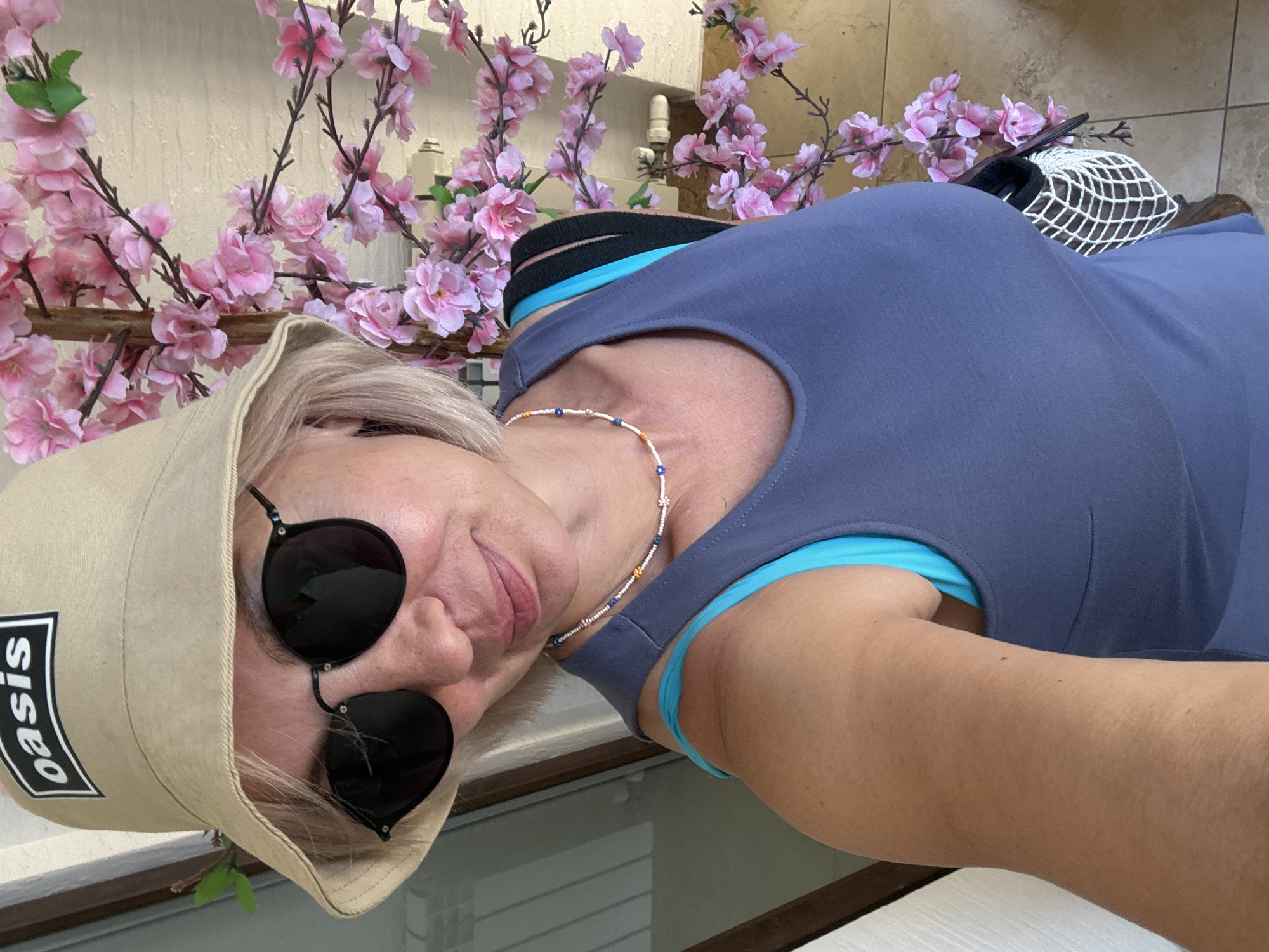 A woman stands indoors, partially framed by a decorative tree with pink flowers. She is wearing a light-colored bucket hat with the word "oasis" in a black rectangle on the front, dark sunglasses, and a blue tank top over a turquoise bra. She has a beaded necklace on. A white netted bag is visible on her right side. The background includes a door on the left and a tiled floor. The location is the city of Sirius, Russia.
