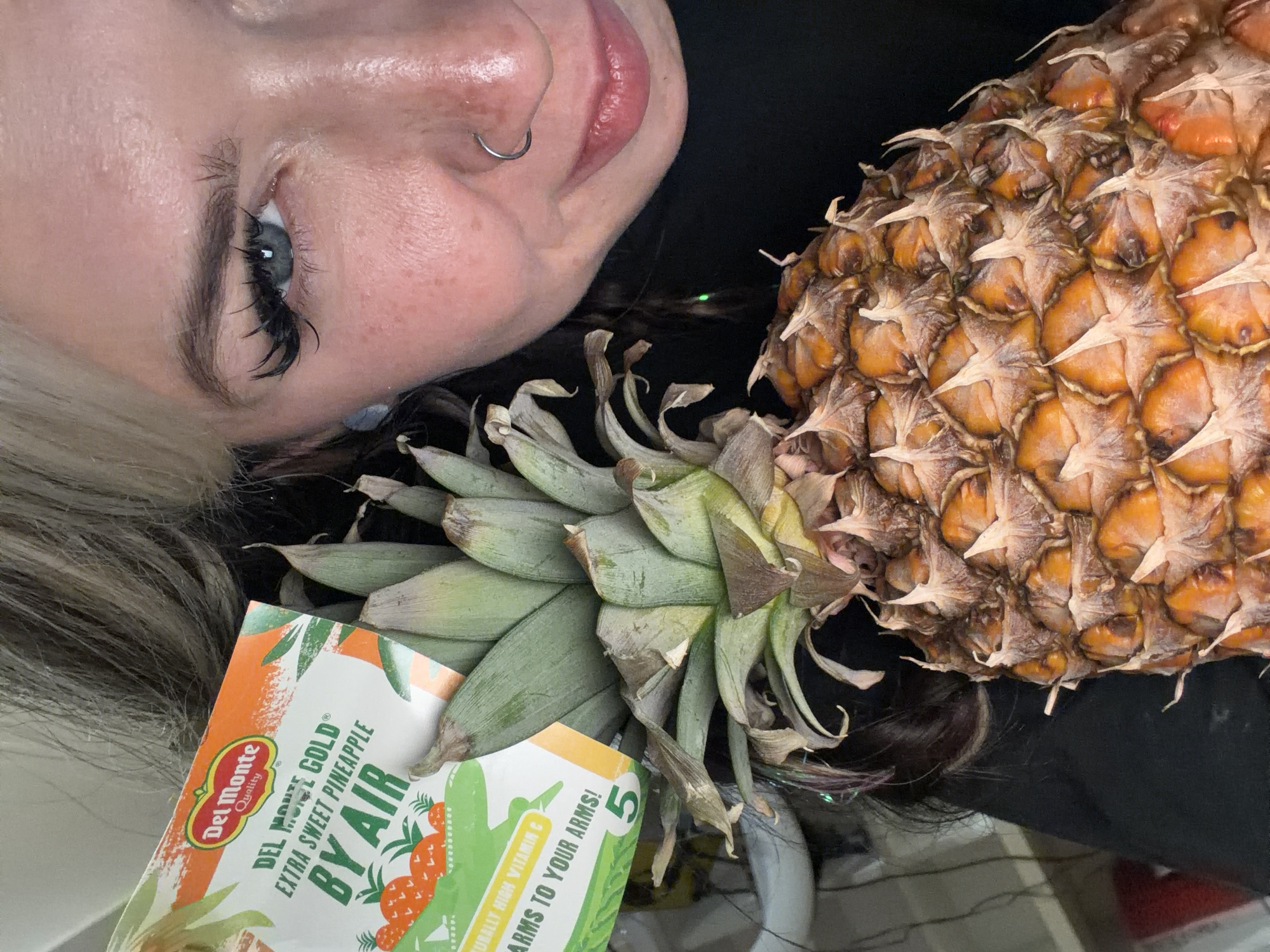 The image features a woman with blonde hair, heavy eyelashes, and a nose ring. She is positioned close to a pineapple, with a label indicating it is a "Del Monte Gold Extra Sweet Pineapple BY AIR". The label also states "Naturally high vitamin C, 5 arms to your arms!". The background is unclear, but what is visible suggests an indoor setting. No specific location in Budva, Montenegro, is identifiable. The interaction is simply a pose, and there are no other people present.