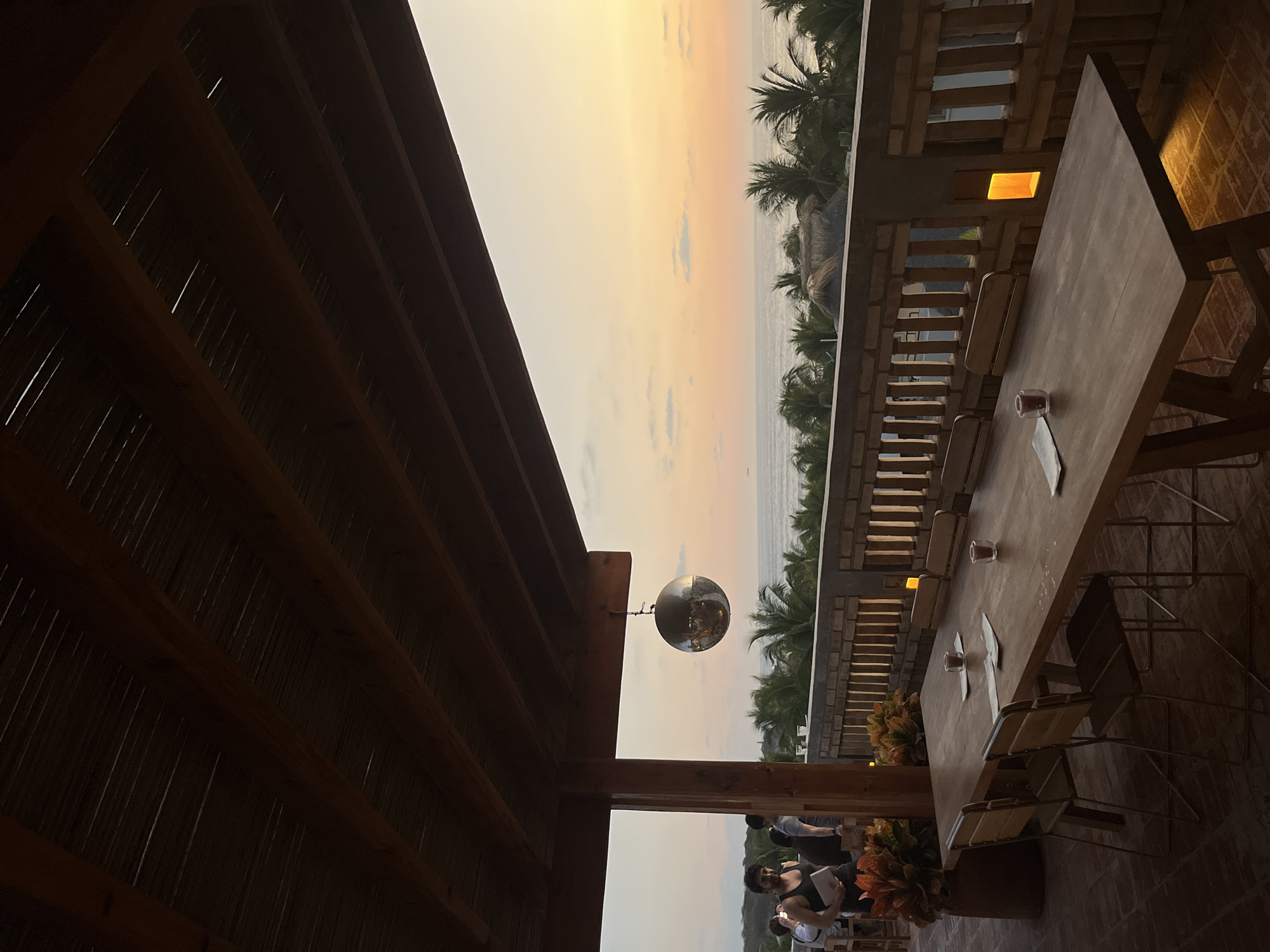 A horizontal, medium-distance shot depicts an outdoor setting with a table, chairs, and an ocean view at dusk in Brisas de Zicatela, Mexico.
To the left, a wooden structure provides a roof over a portion of the scene. Hanging from the structure is a spherical, mirrored object.
In the distance, an ocean and a strip of beach are visible under a yellow and orange sky. Palm trees line the beach.
To the right, a long wooden table with several chairs is set up, with two glasses and napkins on it. Behind the table, a low brick wall with window-like openings runs horizontally. A warm light emanates from one of the openings. At the edge of the frame, there are visible stone-like tiles.
At the bottom of the frame, several people stand near the edge of the structure, with some carrying objects and appearing to be interacting. A potted plant is visible at the bottom.