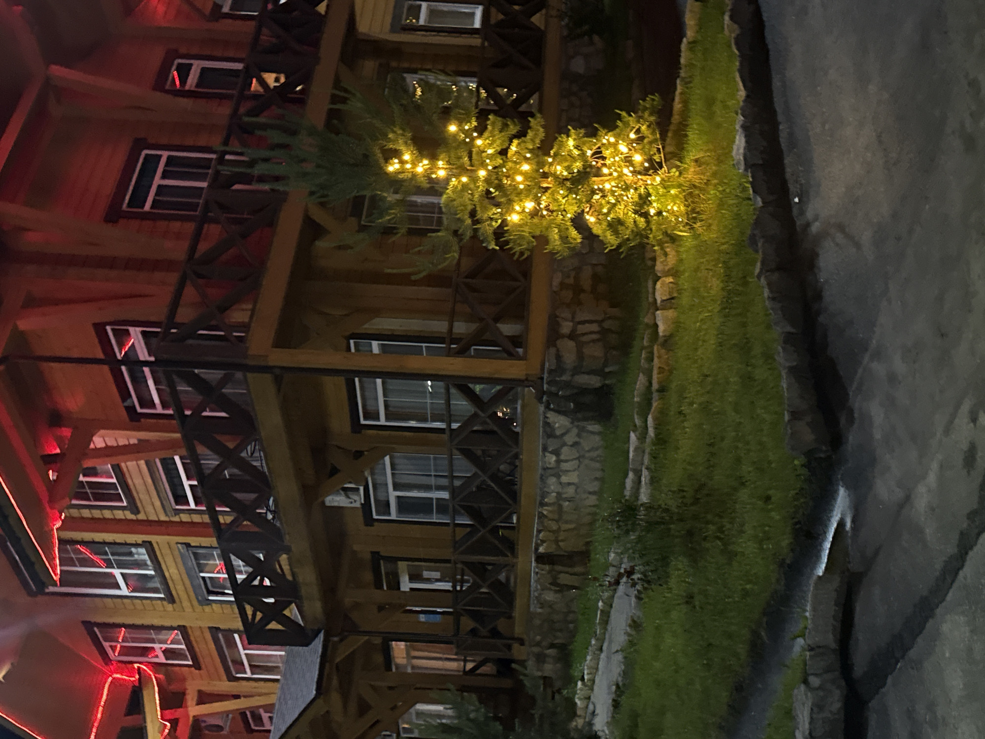 The image depicts a wooden building, possibly a hotel or lodge, with red decorative lights along its roofline. A small evergreen tree in the foreground is adorned with yellow fairy lights. The building has multiple stories with windows and a wooden balcony with a criss-cross railing. The exterior is constructed of dark wooden planks, and the foundation appears to be made of stone. In front of the building, there is a lawn area bordered by stone and a paved walkway or road. The scene is lit by artificial lights, suggesting it is nighttime or dusk. The location is identified as Syrostan, Russia. No people are visible in the image.