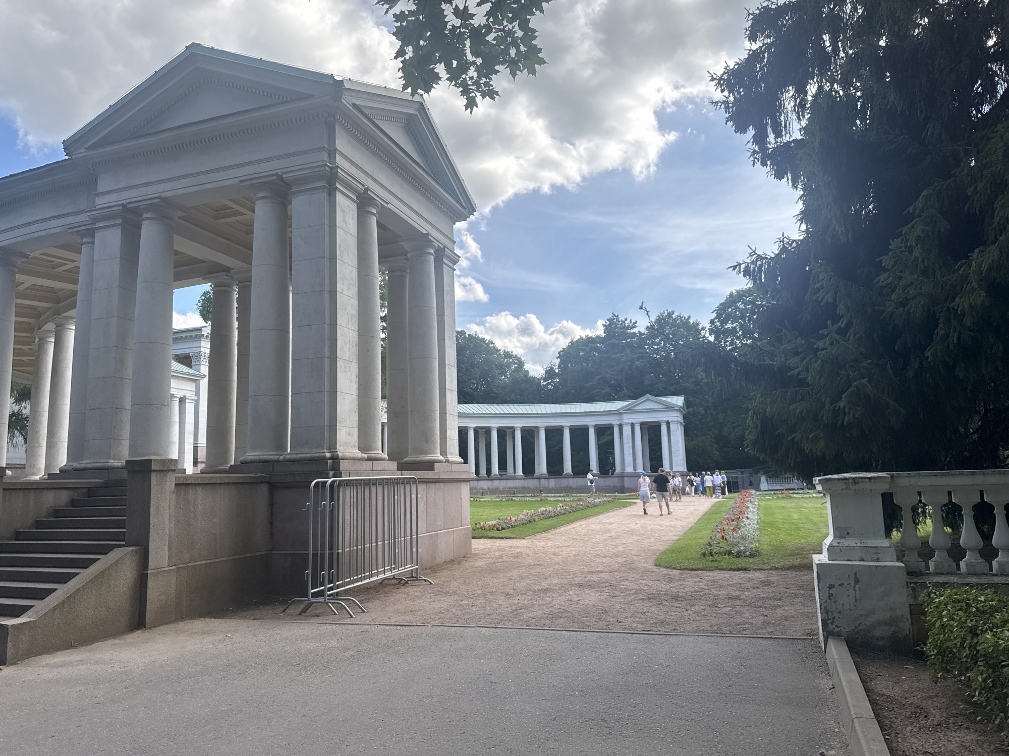 This image depicts a neoclassical colonnade and a building with a triangular pediment in the grounds of the village of dacha housing "Arkhangelskoye", Russia. The foreground features a paved pathway and a gravel path leading towards the colonnade. A row of metal barriers is positioned in front of the steps ascending to the colonnade. Several people are walking along the gravel path and in the grassy area to the right, which is lined with flower beds. The architecture is characterized by stone columns and a white facade. Lush green trees, including a large, dark coniferous tree on the right, frame the scene under a partly cloudy sky.