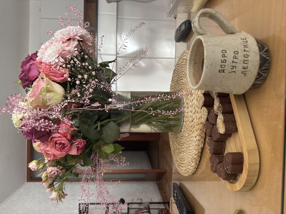 The scene depicts an indoor still life, located in Miljevci, Montenegro. A prominent floral bouquet, comprised of various pink, white, and maroon roses, light pink carnations, and delicate pink filler flowers, stands in a clear glass cylindrical vase, approximately half-filled with water. The vase rests centrally on a circular woven placemat on a glossy, light-brown wooden table. To the left of the bouquet, a black multi-button remote control is present. To the right, a light-colored, speckled ceramic mug with a handle contains a dark beverage and features Cyrillic text: "ДОБРО ЈУТРО ЛЕПОТИЦЕ" (Good Morning, Beautiful), along with a patterned black and white base. Adjacent to the mug, an oval bamboo tray holds approximately 15-18 heart-shaped chocolates, some plain and some topped with a light granular substance. A second black remote control is partially visible behind the mug, and a clear plastic container with yellow/white contents is at the far right edge of the table. The background reveals a light-colored wall to the left, an open wooden doorway leading into another room with a tiled floor, and white kitchen cabinets or furniture on the right. No individuals are present, and no active interactions are depicted.