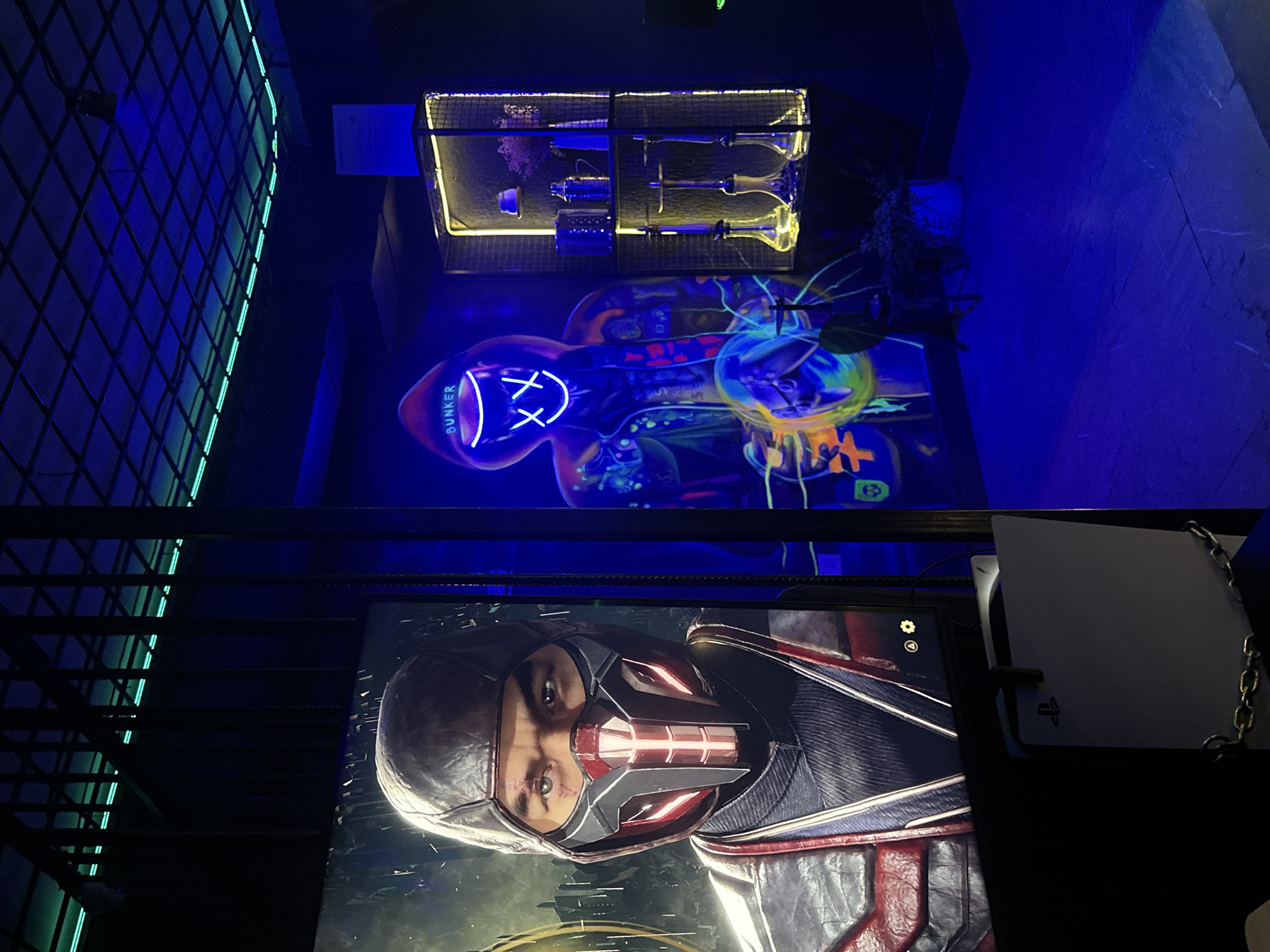 The image presents an indoor setting, likely a gaming room or entertainment area, in Yerevan, Armenia. The scene is illuminated with vibrant blue and yellow lights, creating a futuristic and slightly surreal atmosphere. A large screen displays a close-up of a character with a mask. A PlayStation 5 is visible on the right side of the screen. Above the screen, there is an image with the word "BUNKER" and other illuminated figures. Above that is a display case with glasses. The scene is characterized by neon lights and modern design elements, pointing to a contemporary entertainment space.