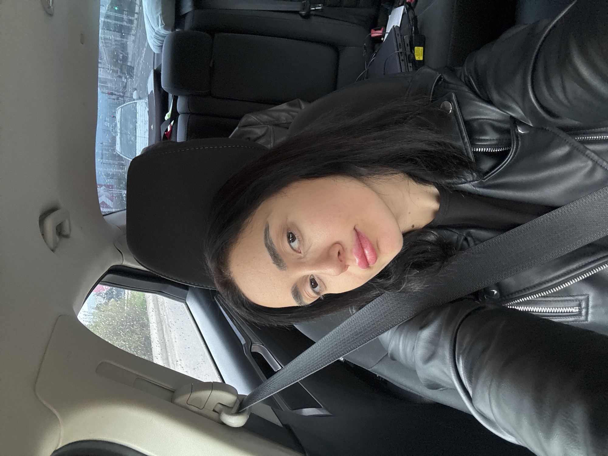 A woman with dark hair and light skin is sitting in the passenger seat of a car, wearing a black leather jacket and a seatbelt. The car is likely stationary due to the raindrops on the window, obscuring the view of the city outside. The cityscape is visible through the window, suggesting the location might be in Tbilisi, Georgia. The car's interior is dark, with black seats.