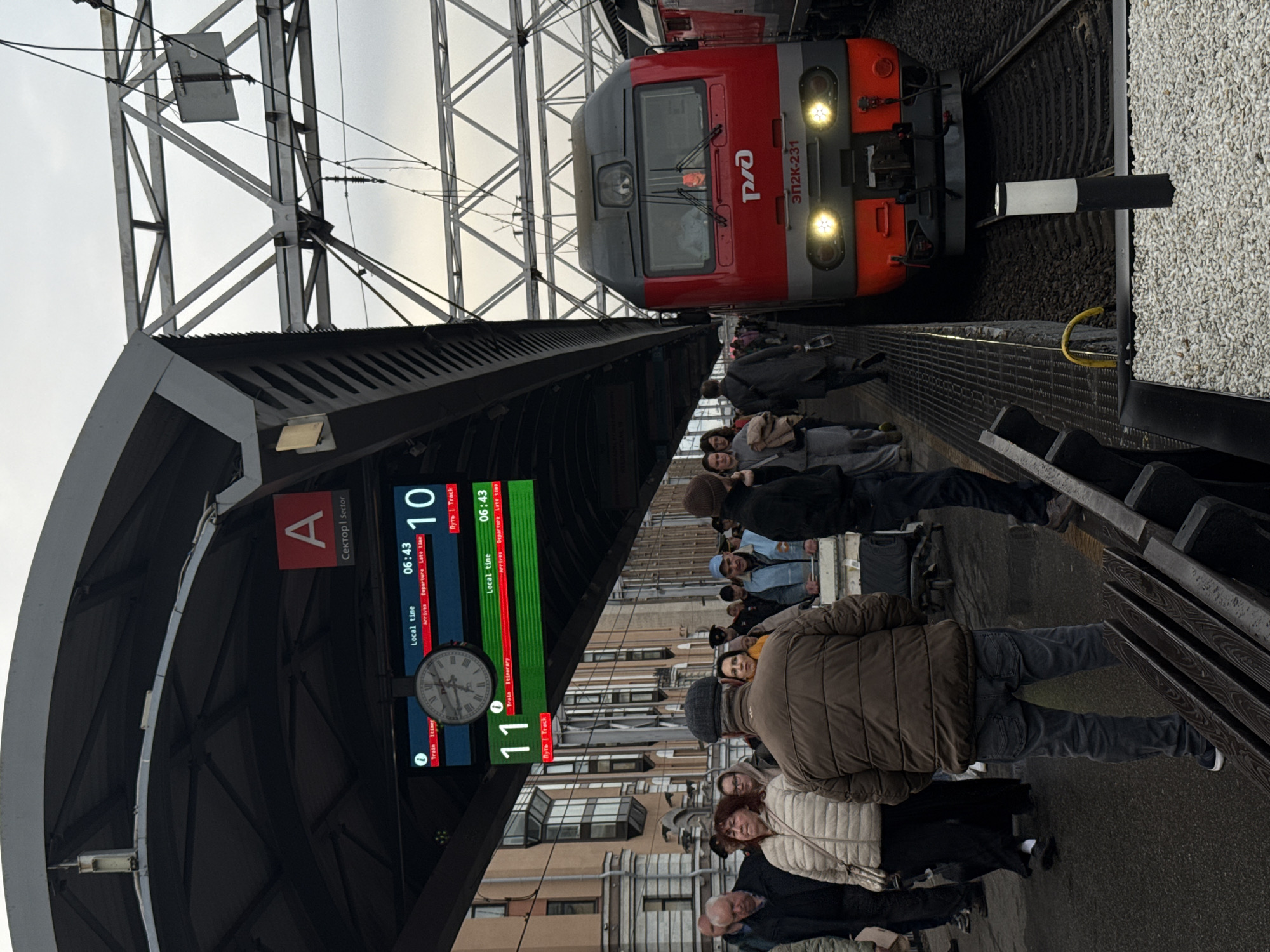 The image depicts a train station platform with a red and gray train approaching the station, likely in Saint Petersburg, Russia, indicated by the Russian Railways logo and Cyrillic script on the train (ЭП2К-231). The platform is crowded with people waiting for or disembarking from the train. Above the platform, there are digital displays showing train arrival and departure information, including a clock showing the local time and track numbers. The displays read "Local time 06:43" for both Track 10 and Track 11. There's a sign indicating "Sector A." Overhead structural elements of the station are visible, including supporting beams and wiring.
