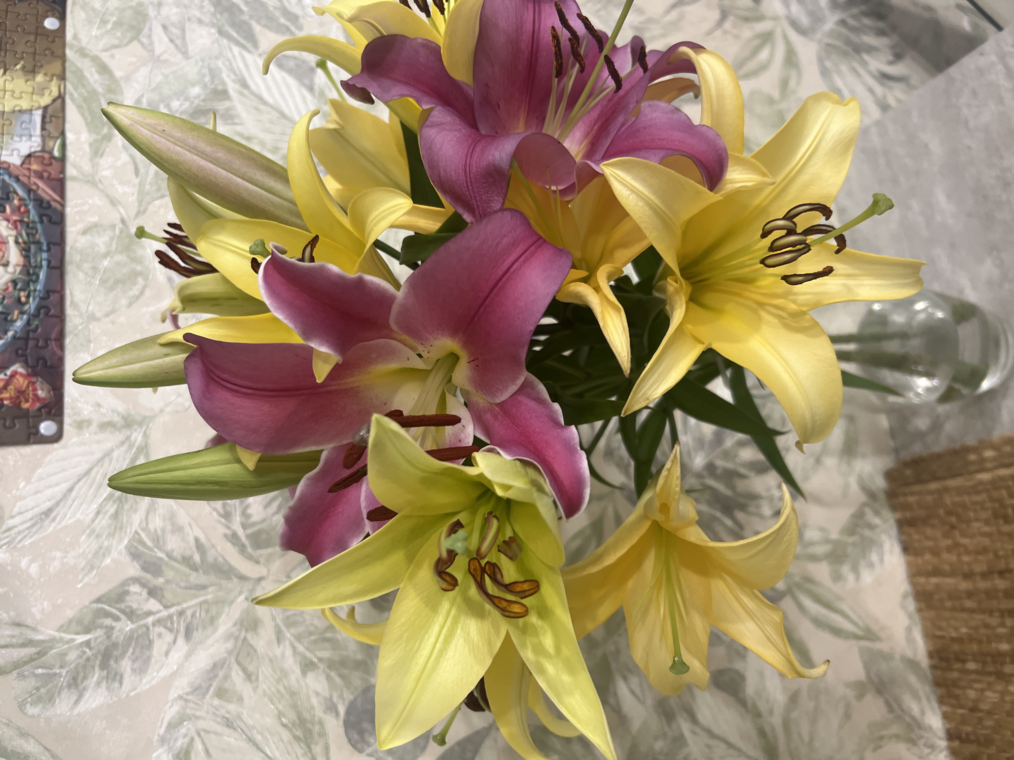A bouquet of yellow and pink lilies is displayed in a clear glass vase on a table. The background features a wall with a floral wallpaper pattern. A puzzle is visible on the wall above the flowers. The lighting suggests an indoor setting. No specific location in Volgograd, Russia, can be determined from the image.
