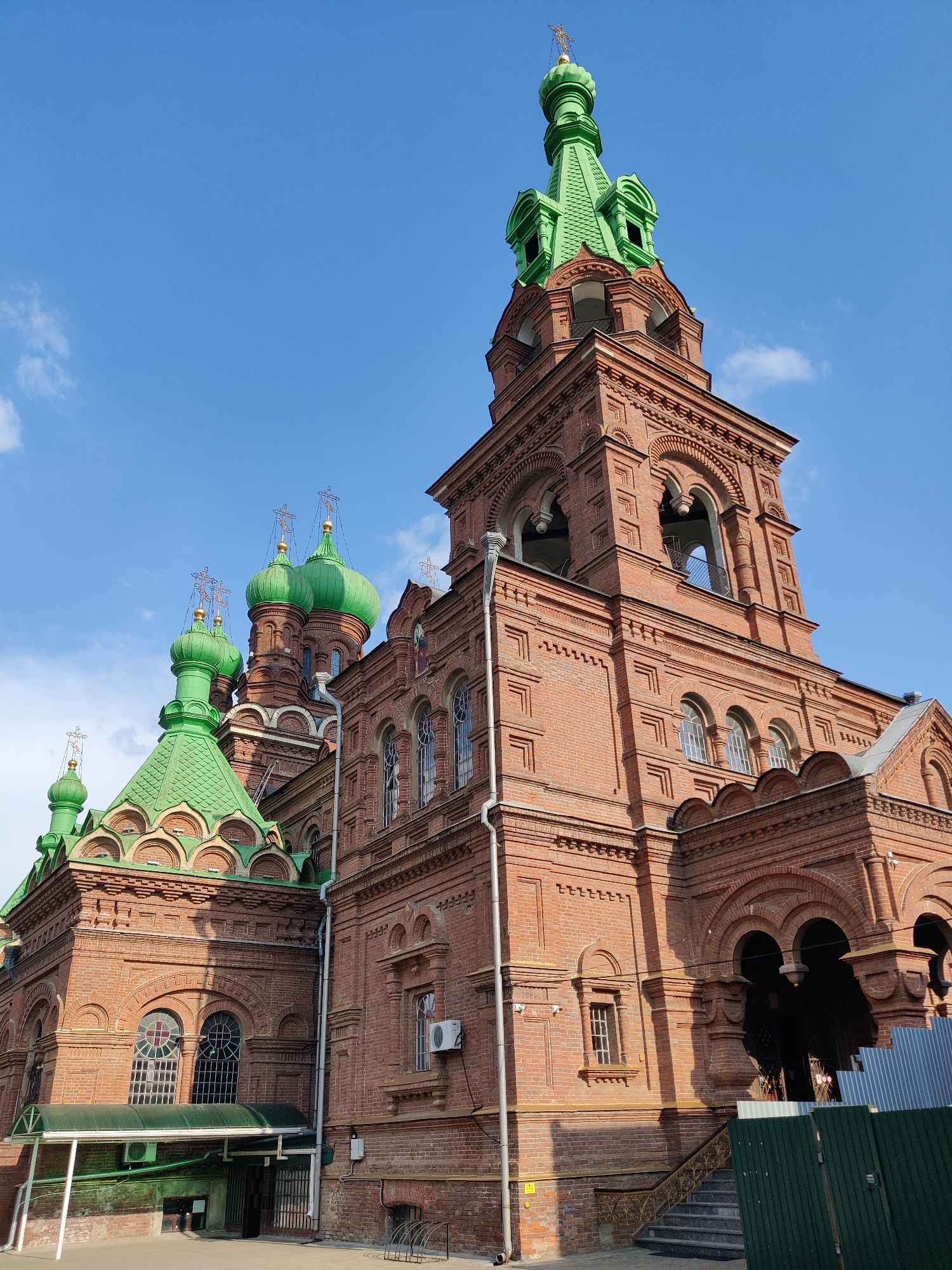 The image shows the exterior of the Catherine Cathedral in Krasnodar, Russia, on a clear day with a few scattered clouds. The cathedral is constructed of red brick and features multiple green domes topped with crosses. A prominent bell tower with arched openings is visible on the right side of the image. The architectural style is characteristic of Russian Orthodox churches. No people are visible in the frame. The foreground shows a paved area and some fencing.