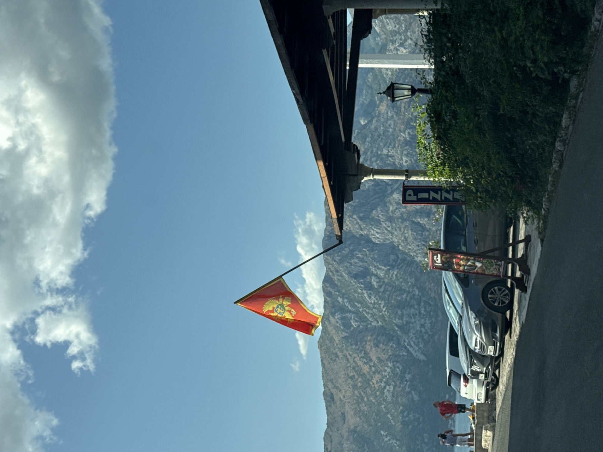 The image depicts a street scene in Prčanj, Montenegro, with a focus on a building featuring a covered outdoor seating area and a flag of Montenegro prominently displayed. The flag, red with a gold double-headed eagle crest, is attached to a pole extending from the building's roofline. In the background, a rugged mountain range dominates the landscape under a blue sky with scattered clouds.

In the foreground, parked cars are visible along the roadside. A sign with the word "PIZZA" in vertical lettering is positioned near the cars, suggesting a restaurant. The building itself appears to be a restaurant or cafe, with architectural elements like columns supporting the roof. Lush green bushes flank the building and the seating area.

Several people are present in the scene. On the left, two individuals are standing near the water's edge, facing away from the camera. Closer to the cars, two men are standing. One is wearing a red t-shirt, and the other appears to be wearing a light-colored shirt. The overall environment suggests a warm, sunny day in a coastal town, likely during the tourist season. The road in the foreground is asphalt.