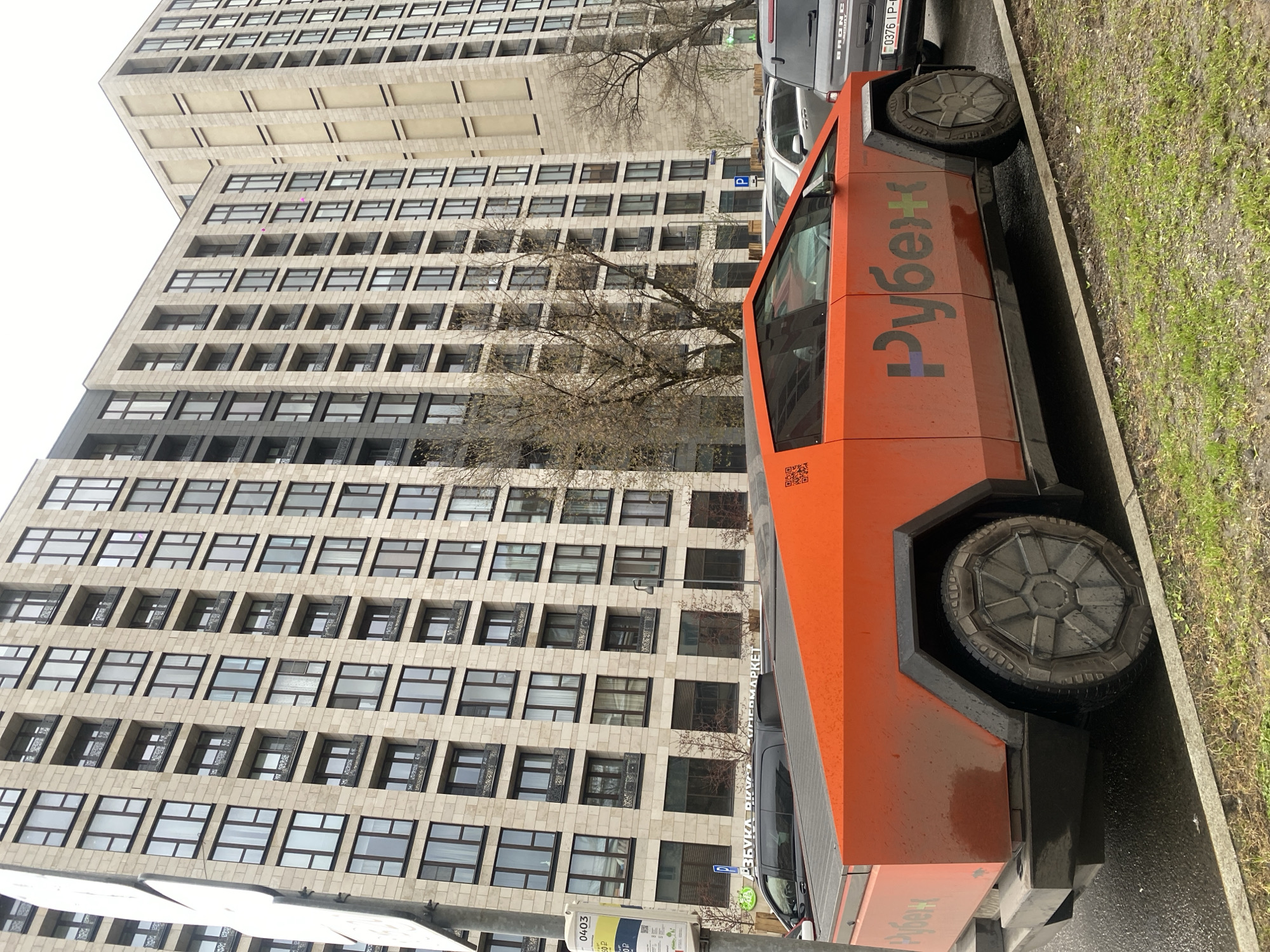 The image shows a bright orange, angular vehicle parked on a street in Moscow, Russia. The vehicle has the word "Рубеж" (Rubezh) written on its side in Russian, with the letters colored in a gradient of purple, blue, and green. It has unusual, disc-shaped wheels. The vehicle appears to be of a futuristic or unconventional design.
Behind the vehicle, a tall, multi-story building with a repetitive pattern of windows dominates the background. A tree with bare branches is visible in front of the building. To the side of the orange vehicle, other cars are parked, including what appears to be a silver or grey Ford Bronco. A bus stop sign is also visible near the curb. The street is adjacent to a grassy area. The lighting suggests an overcast day.