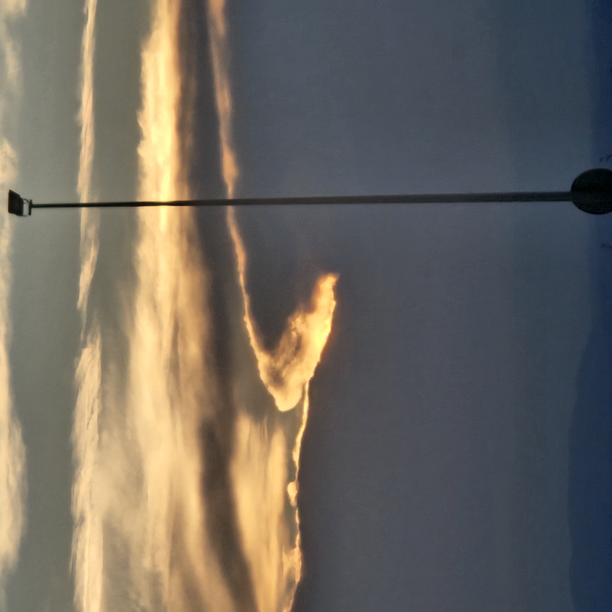 The image depicts a sunset sky in Algora, Spain. The sky is divided into two main areas: a section with golden, illuminated clouds on the left, and a darker, blue-grey section on the right. A prominent cloud formation in the center resembles an abstract shape, highlighted by the setting sun. In the foreground, a straight, horizontal pole (likely a streetlight) extends across the entire width of the image, intersecting the sky. On the left end of the pole, a streetlight fixture is visible, while the other end culminates in a circular lamp head. No people are visible in the image.
