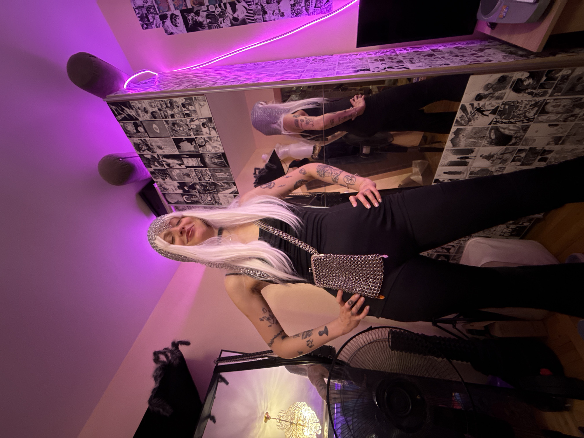An individual stands in a room with purple lighting, facing a full-length mirror. The person is wearing a black sleeveless jumpsuit and a silver chainmail-like hood. They have long, straight blonde hair and a chainmail purse hanging from their shoulder. Their hands are on their hips. The mirror reflects the individual and the room behind them, which is decorated with numerous black and white manga panels. In the foreground, a black fan is visible on the left side of the image, and a chandelier hangs in the reflection in the mirror. The background features a large mirror with manga panel decorations on its frame and wall. The overall impression is of a personal space, possibly a bedroom or dressing area, with a distinct aesthetic. The location is not specified as Moscow, Russia.