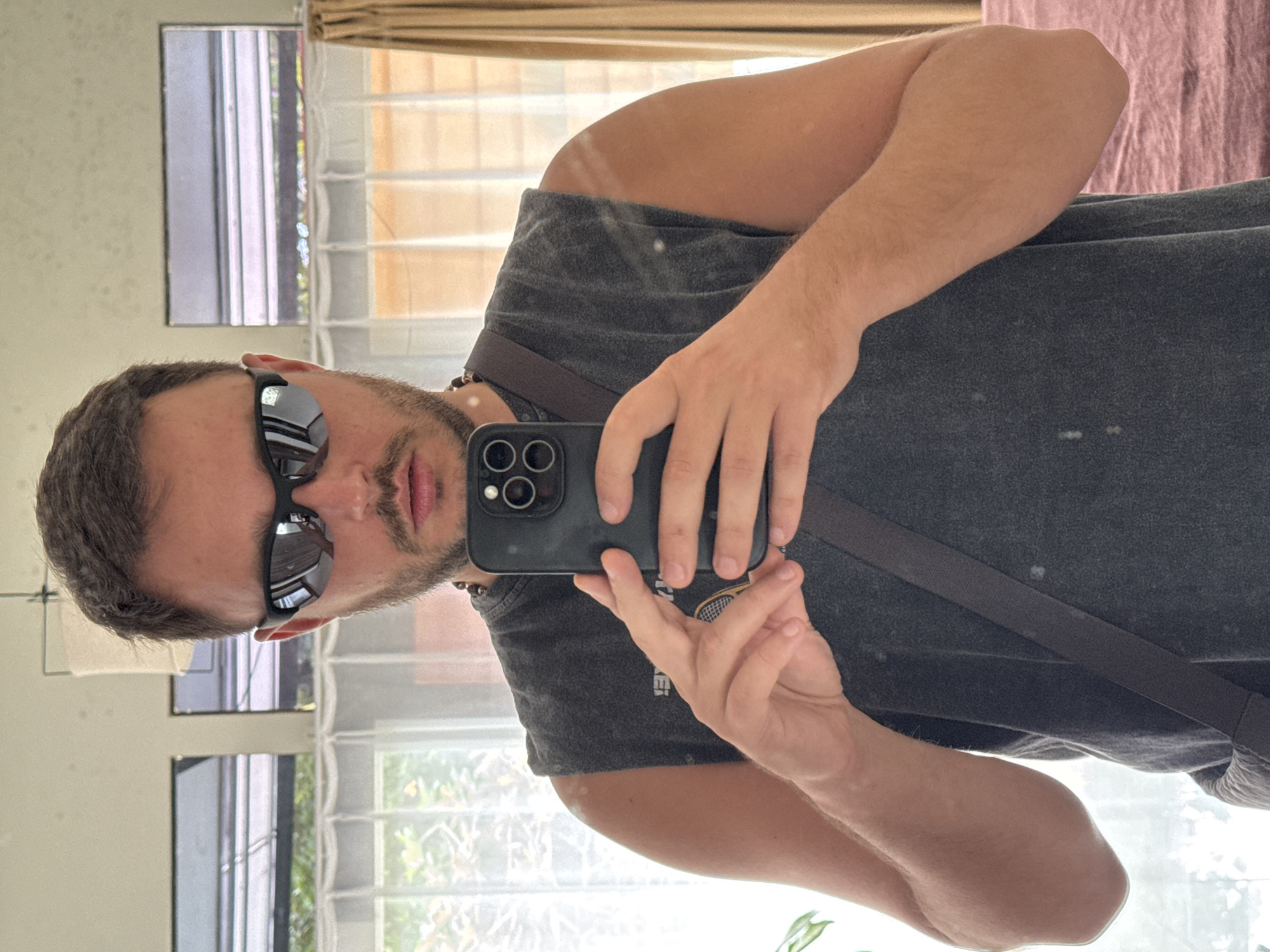 A man with short brown hair and a goatee is taking a selfie in a mirror. He is wearing sunglasses and a gray t-shirt with a strap crossing over his shoulder. He is holding a black smartphone with a triple-lens camera. The background includes a window with white curtains and a glimpse of greenery outside. The location is Kuta Selatan, Indonesia.