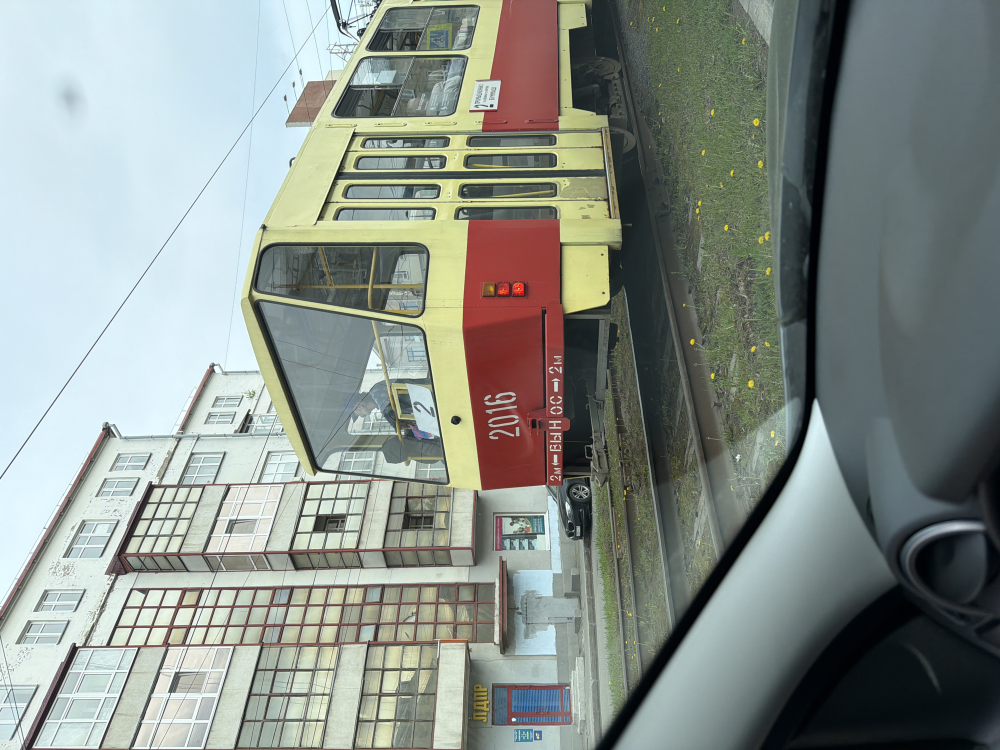 The image shows a view from inside a car, looking out at a red and yellow tram traveling alongside a building. The building is white with numerous windows, some of which are bordered with red. The tram is numbered "2016" and has Russian text on the side. A grassy area with yellow flowers is visible beside the tram tracks. Another vehicle is parked on the street. The location is Unknown, Unknown.