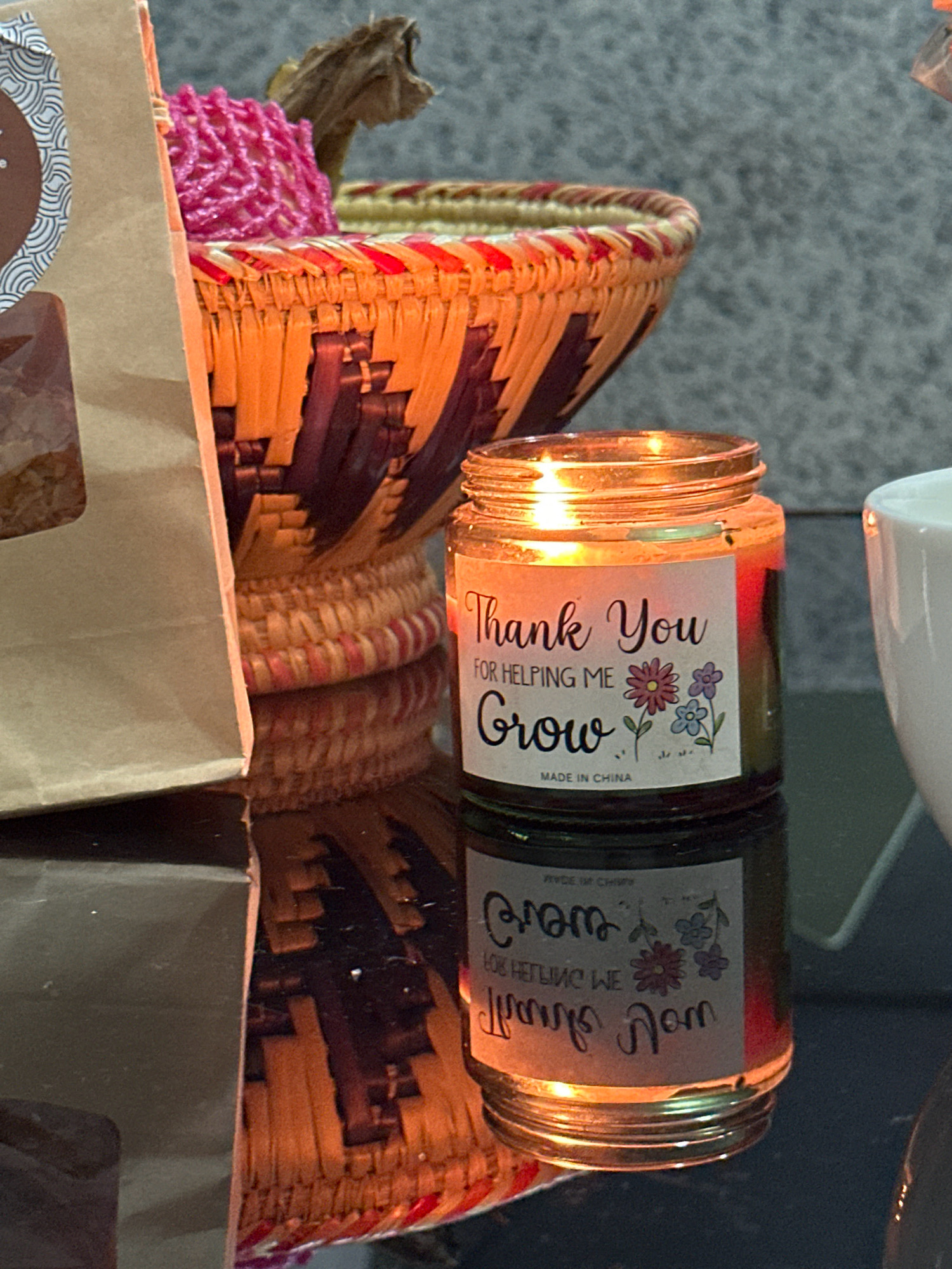 A lit candle in a glass jar with a label that reads "Thank You for Helping Me Grow" sits on a reflective dark surface. The candle is a warm orange and yellow, with a flame at the top. The label also features small floral illustrations. To the left of the candle is a brown paper bag, partially obscuring a woven basket with orange and dark purple detailing. The basket contains a bright pink, textured item. The background is a textured gray wall. The scene appears to be indoors, possibly on a table or shelf, in an environment that suggests it might be in the city of Lalitpur, Nepal, given the decorative style of the basket and the general presentation. There are no people visible in the image. The lighting is warm, likely from the candle itself and an ambient light source.