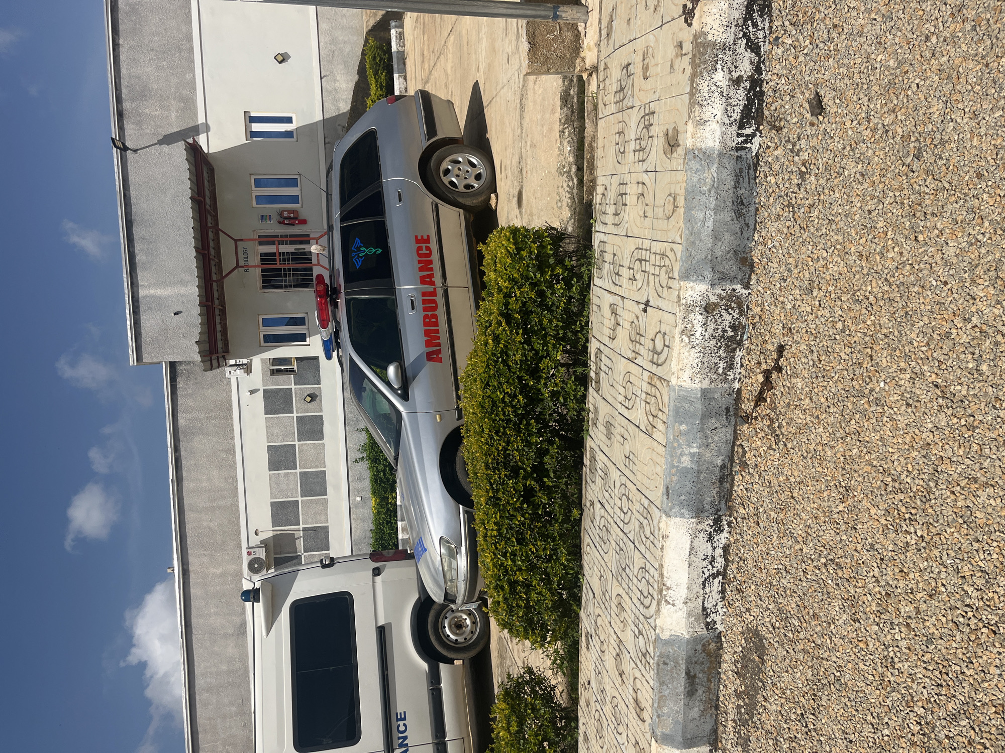 This media file shows an ambulance parked next to a building, likely a hospital or clinic, in Jos, Nigeria. The ambulance is silver with red and blue lights on the roof. The word "AMBULANCE" is written in red lettering on the side of the vehicle. Next to the ambulance is a white van, also likely a medical vehicle, with the word "ANCE" visible in blue lettering on its side. A green bush is planted in front of the ambulance. The building has several windows and a red awning over the entrance. The ground is paved with patterned tiles and bordered by a strip of gravel. The sky is blue with a few white clouds. The scene appears to be set during the daytime, with bright sunlight casting shadows. There are no people visible in the frame. The overall impression is one of a functional medical facility, possibly during a quiet period.