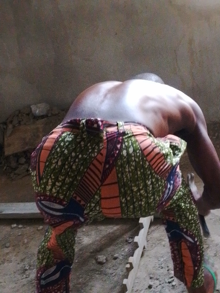 In Enugwu-Ukwu, Nigeria, a shirtless individual is depicted from a rear, slightly elevated perspective, bending forward at the waist. The person, exhibiting a dark skin tone, wears trousers patterned with vibrant green, orange, red, and blue geometric and organic motifs. Their muscular back and shoulders are visible, illuminated by a localized light source creating highlights. The individual's right hand grasps a hammer, identifiable by its dark handle and metallic head. The ground is an uneven surface of dirt and scattered small aggregate. Present on the ground are several pieces of light-colored timber: a long, narrow plank positioned horizontally across the mid-ground, and a smaller, carved piece with an undulating or serrated edge. A portion of a green flip-flop is visible on the individual's left foot, positioned near the bottom right of the frame. The background consists of a plain, light-toned, untextured wall. The scene is consistent with manual labor in a construction or workshop environment.